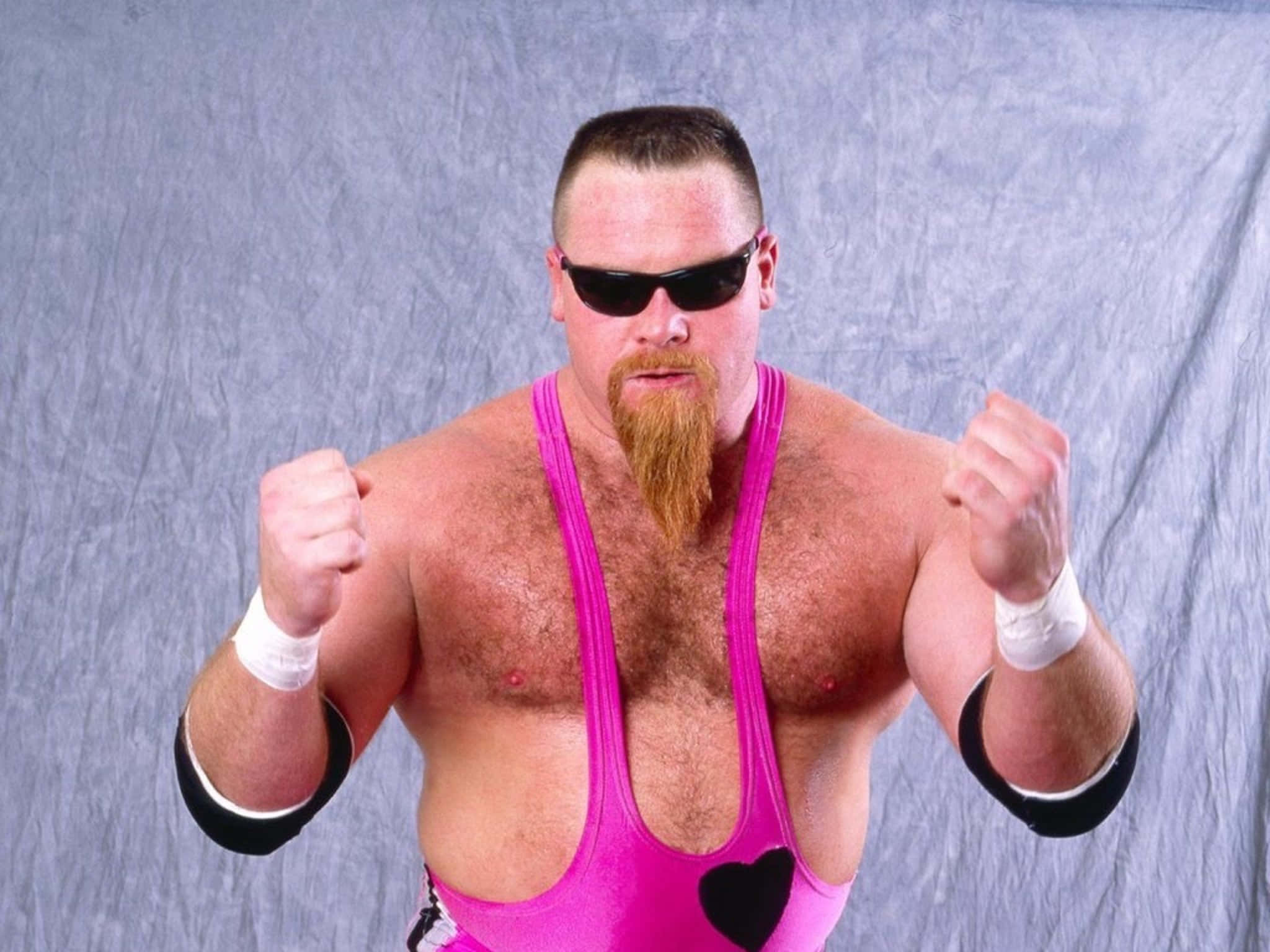 Wrestling 桌布 壁紙 — 一位肌肉發達的摔角手穿著亮粉紅色緊身衣和黑色太陽眼鏡，在灰色背景下舉起拳頭擺姿勢。