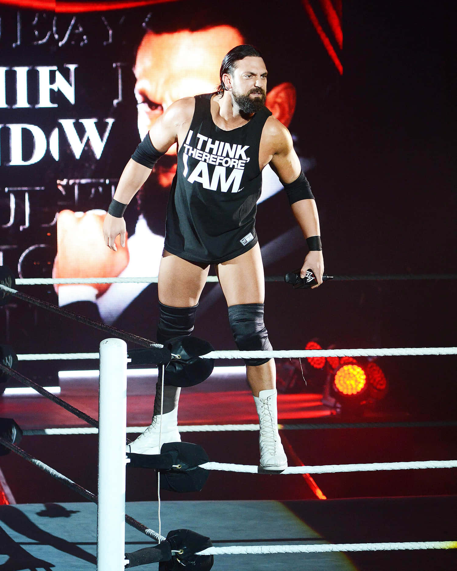 WWE 桌布 壁紙 — 一位肌肉發達的摔角手穿著印有「I THINK THEREFORE I AM」的黑色背心，在明亮的舞台燈光下站在摔角台的角柱上。