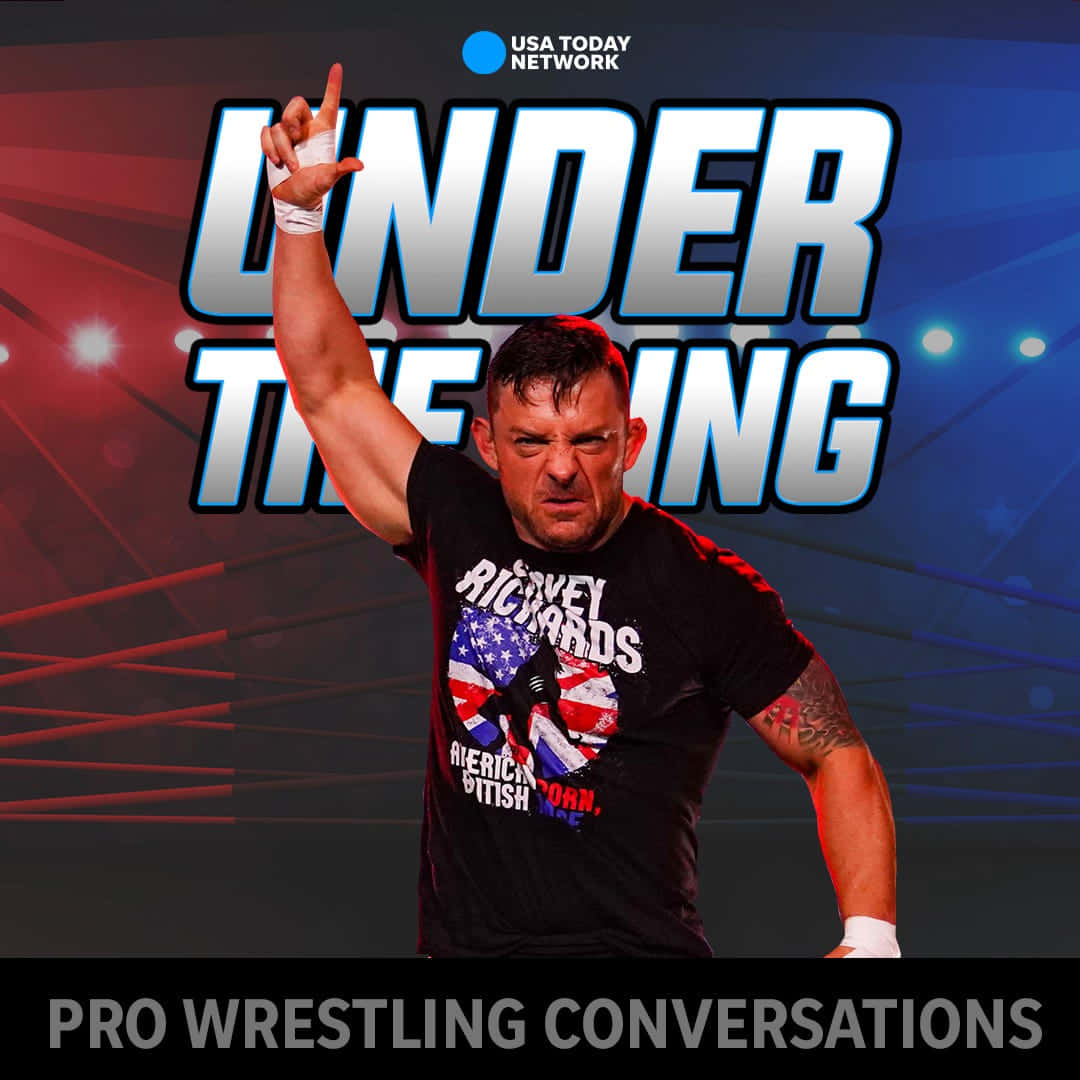 WWE 桌布 壁紙 — 一位肌肉發達的摔角手穿著印有美國和英國國旗圖案的黑色T恤，在明亮的場館燈光下舉起右手擺出勝利姿勢。