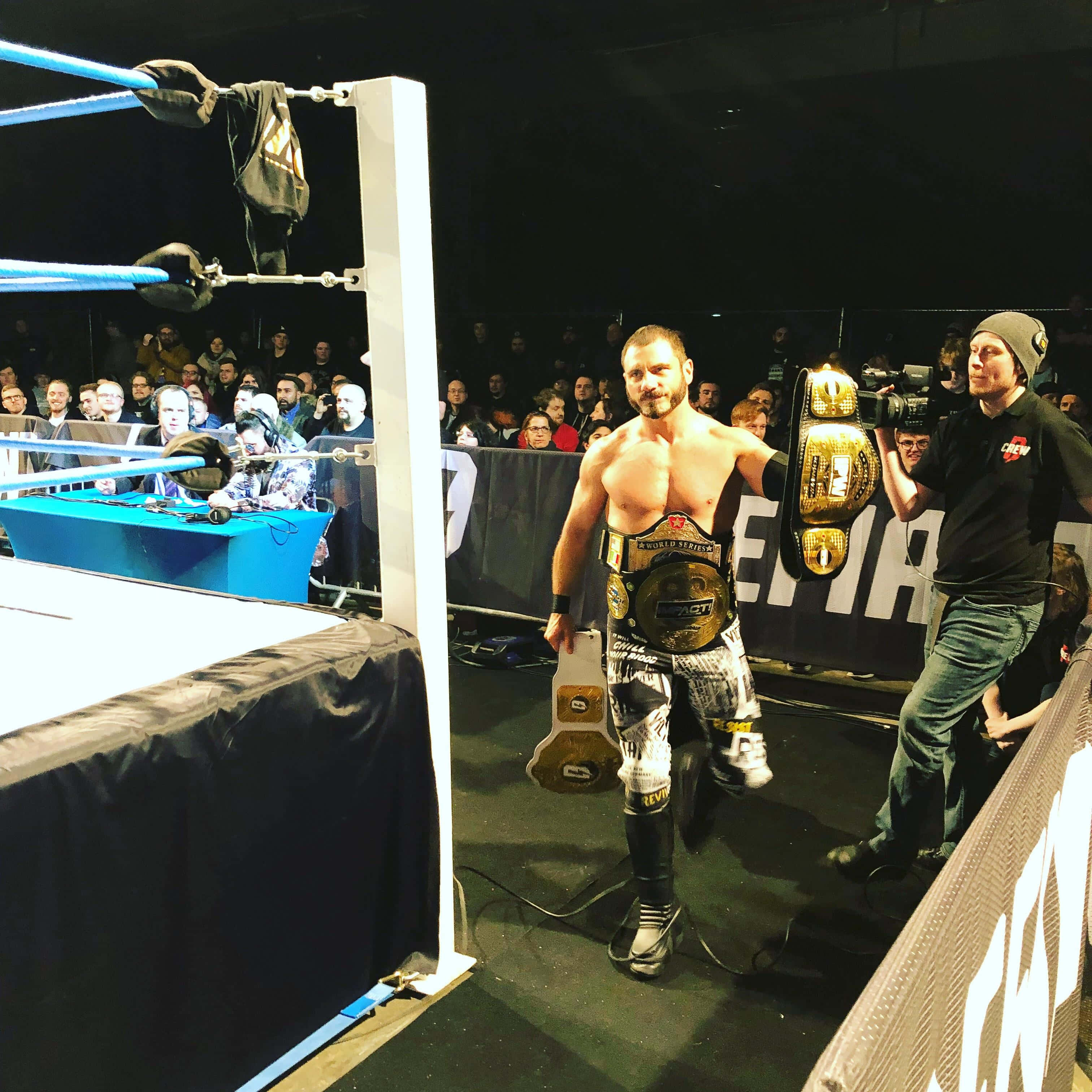 WWE 桌布 壁紙 — 一位肌肉發達的摔角手穿著圖案短褲，在明亮的場館燈光下自信地走出擂台，手持兩條冠軍腰帶。