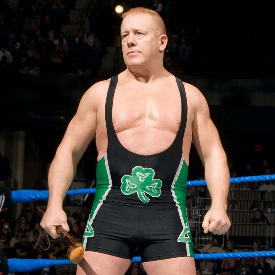 WWE 桌布 壁紙 — 一位肌肉發達的摔角手穿著帶有三葉草圖案的黑色和綠色緊身衣，站在摔角場上手持木棒。