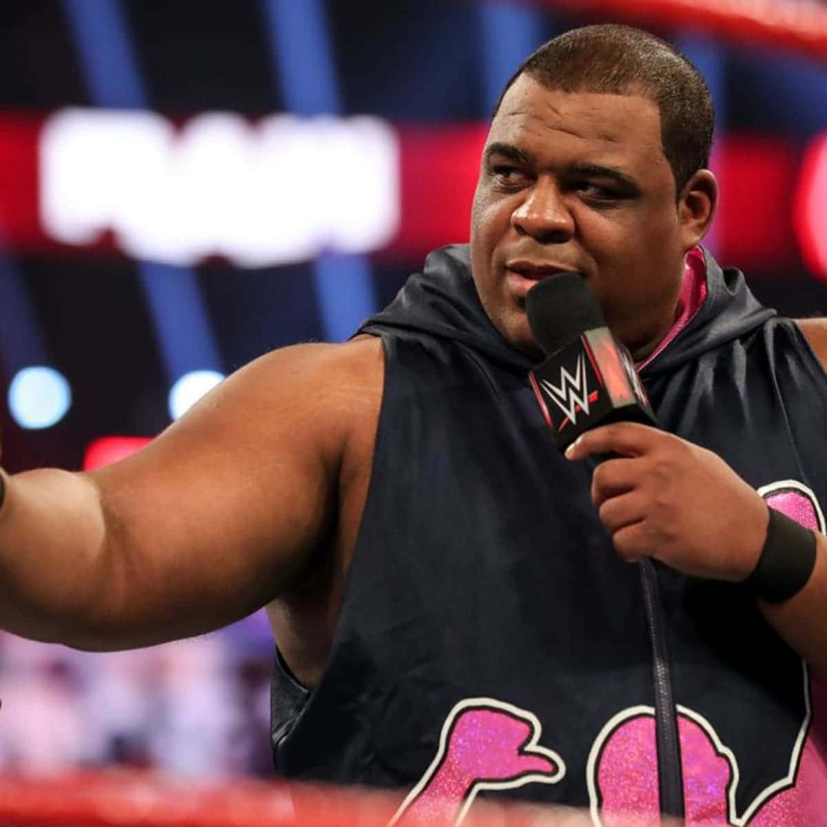 WWE 桌布 壁紙 — 一位肌肉發達的摔角手穿著帶有粉紅色閃粉圖案的黑色連帽衫，手持WWE麥克風，在燈光璀璨的場館中向觀眾做手勢。