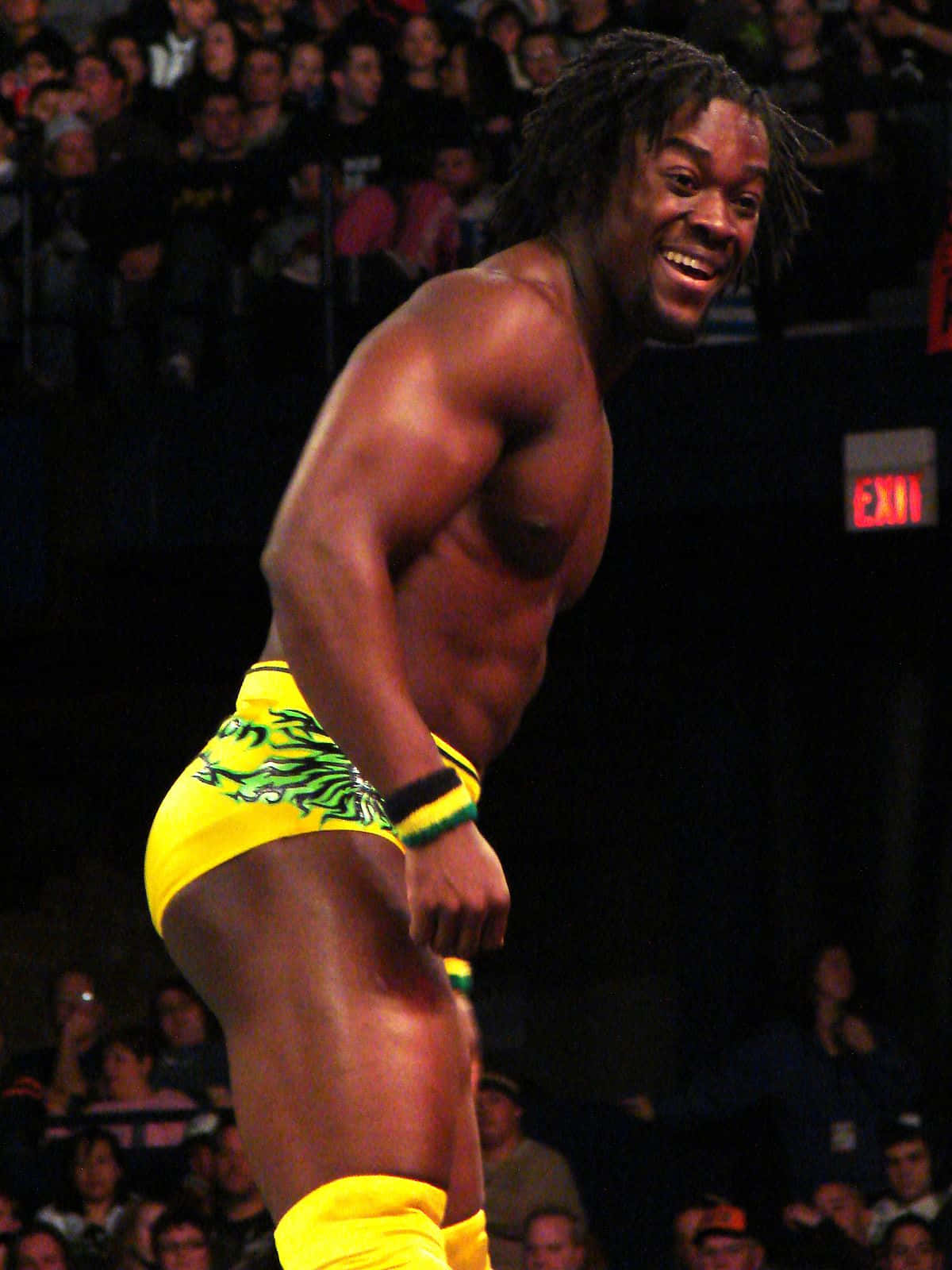 WWE 桌布 壁紙 — 一位肌肉發達的摔角手穿著帶有綠色火焰圖案的亮黃色短褲，在場館燈光下對著歡呼的人群自信微笑。