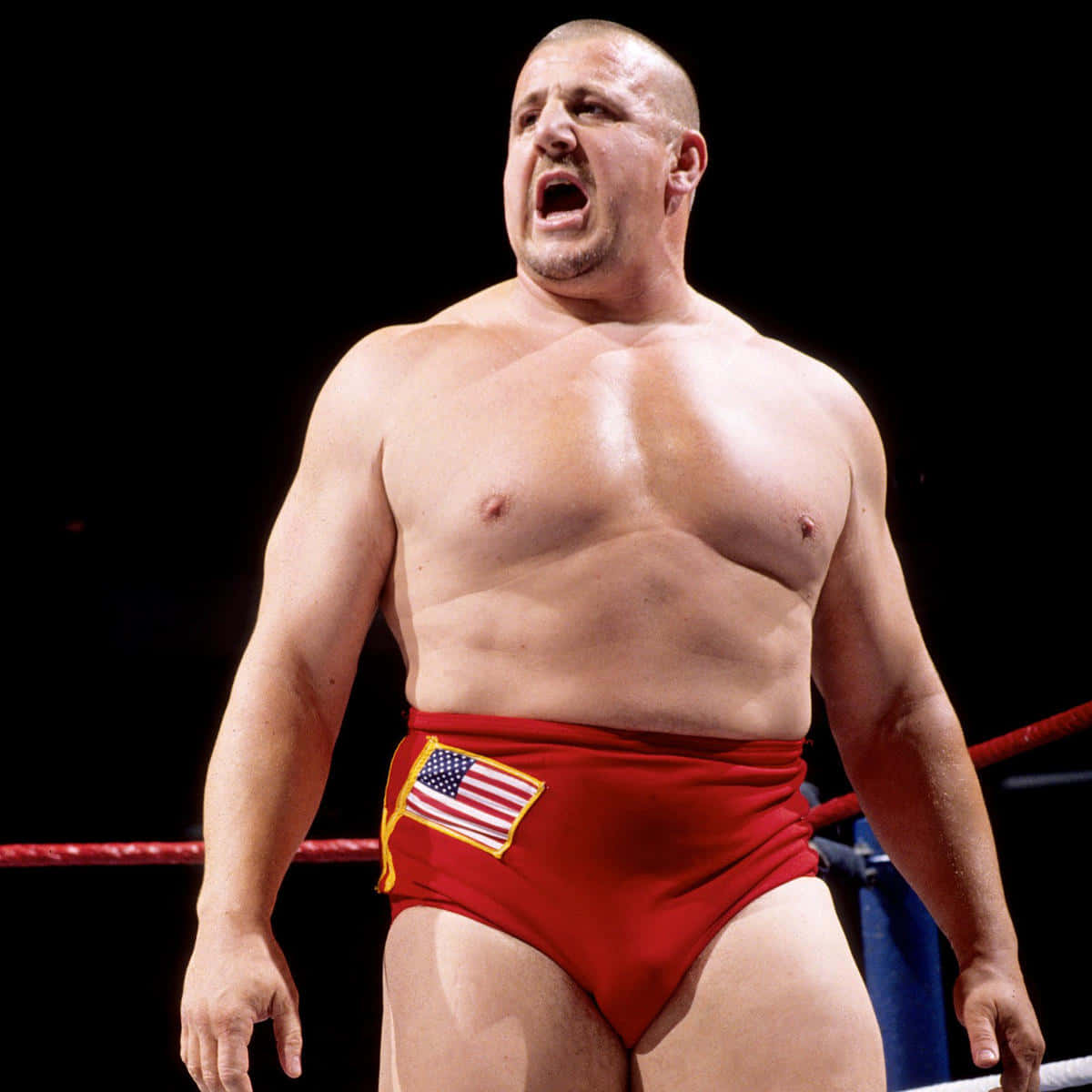 WWE 桌布 壁紙 — 一位肌肉發達的摔角手穿著帶有美國國旗補丁的紅色短褲，在昏暗的擂台上咆哮。
