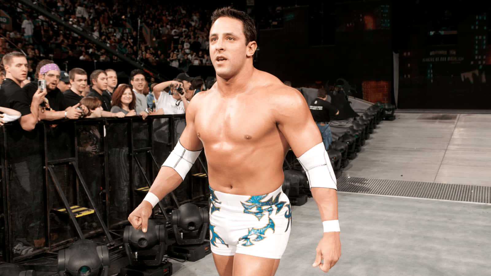 WWE 桌布 壁紙 — 一位肌肉發達的摔角手穿著帶有藍色龍紋的白色緊身褲，自信地沿著斜坡走向歡呼的人群。