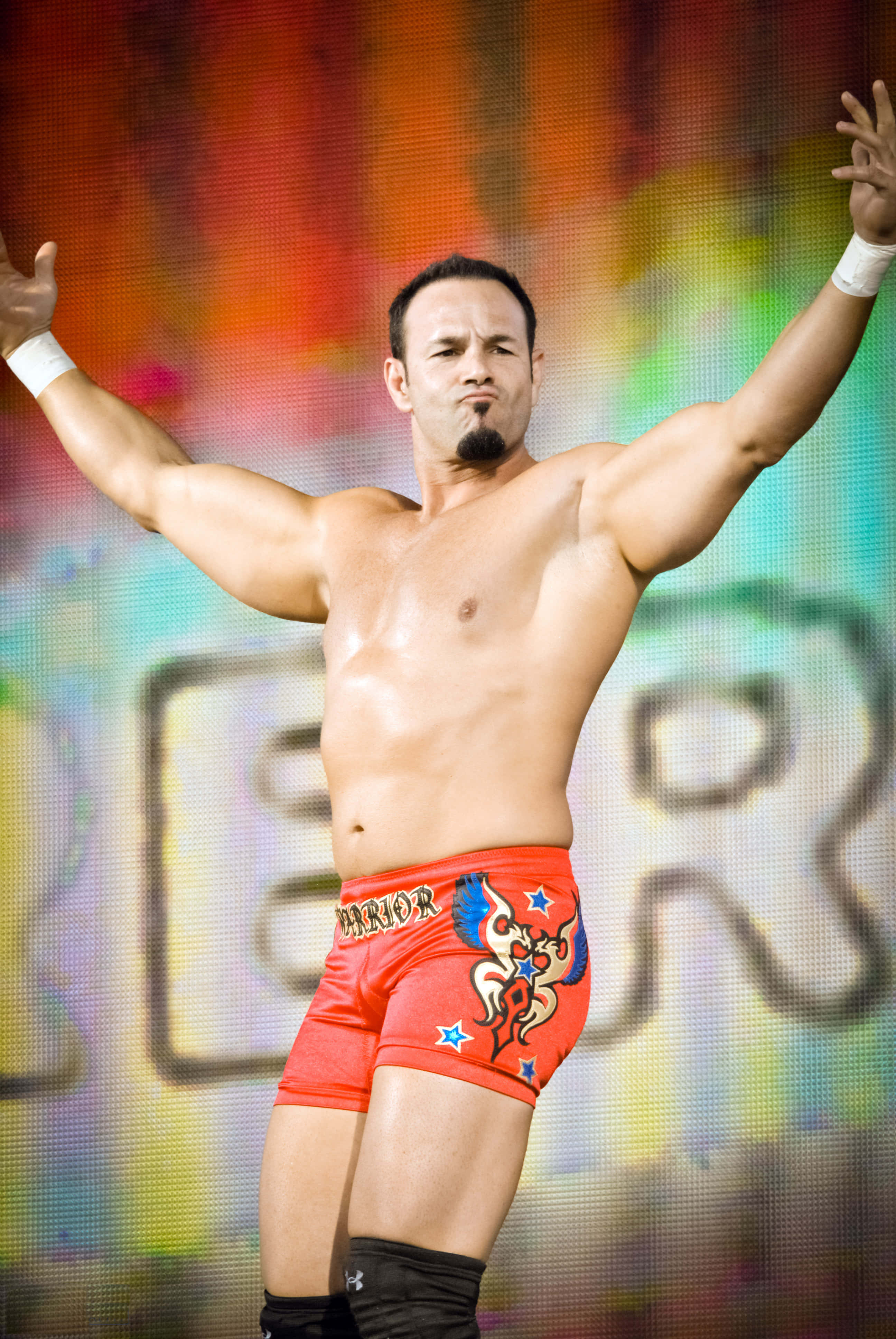 WWE 桌布 壁紙 — 一位肌肉發達的摔角手穿著帶有龍圖案的紅色短褲，在充滿活力的多色LED背景前勝利地舉起雙臂。