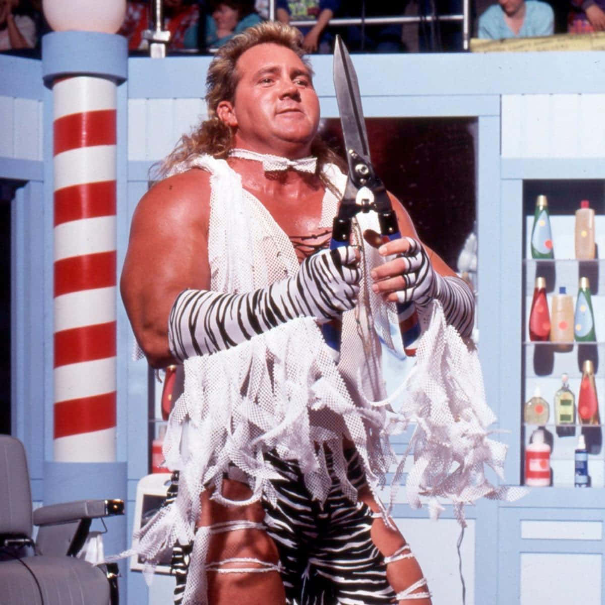 Brutus Beefcake 桌布 壁紙 — 一位肌肉發達的摔角手穿著斑馬紋裝備，手持巨型剪刀，站在色彩繽紛、主題鮮明的摔角場上。