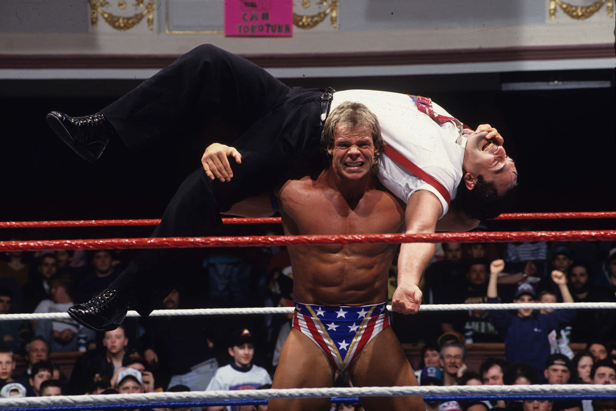 WWE 桌布 壁紙 — 一位肌肉發達的摔角手穿著星條短褲，在明亮的場館燈光下於摔角台上將對手高舉過頭。