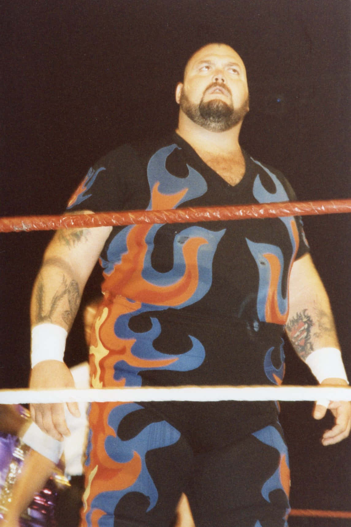 WWE 桌布 壁紙 — 一位肌肉發達的摔角手穿著火焰圖案的黑色服裝，站在有紅色繩索的摔角擂台內，場館燈光璀璨。