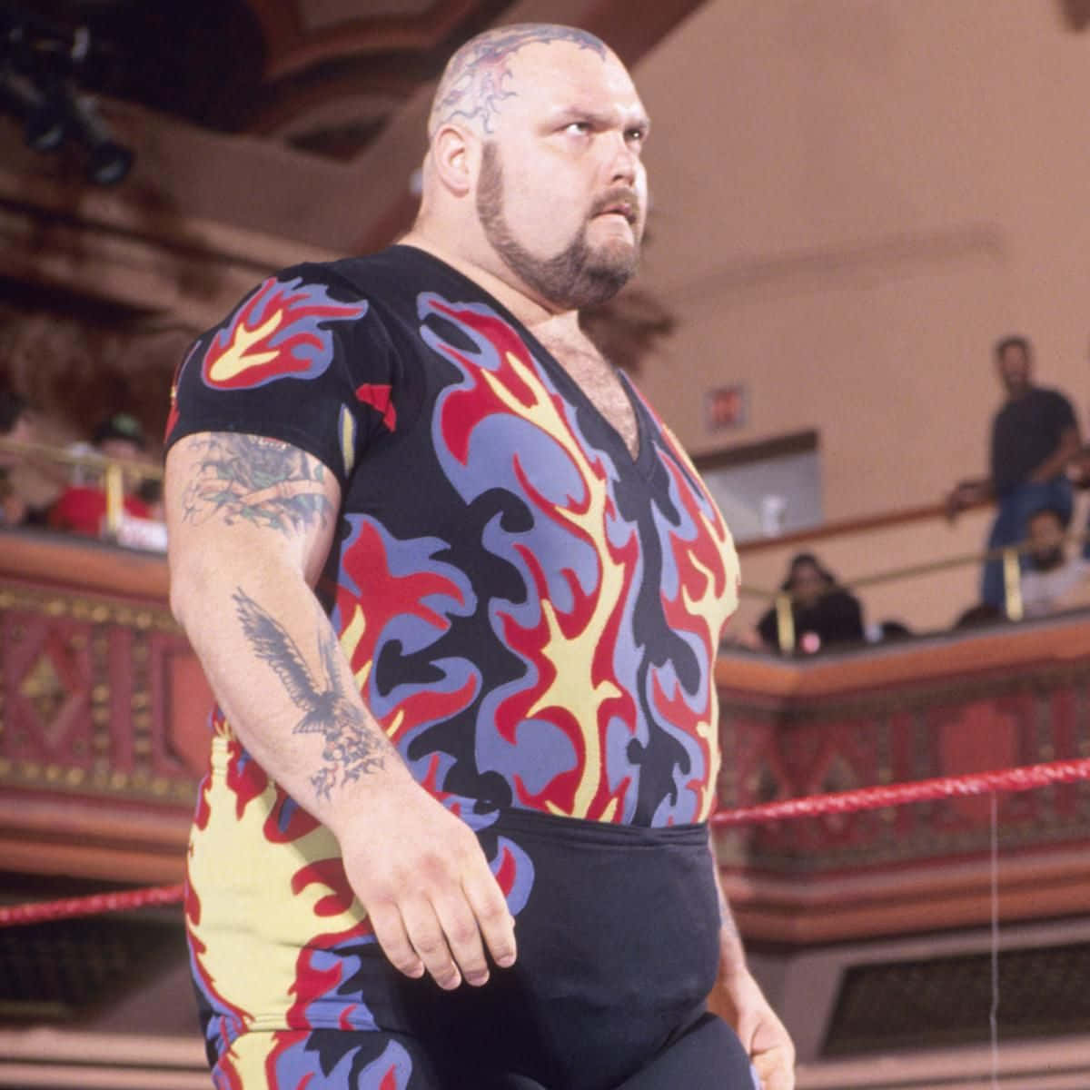 WWE 桌布 壁紙 — 一位肌肉發達的摔角手穿著火焰圖案緊身衣，帶有刺青和光頭，站在擂台上。