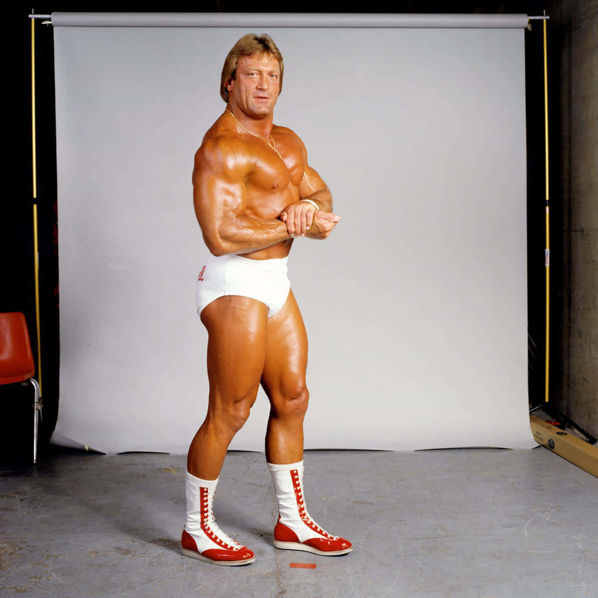 WWE 桌布 壁紙 — 一位肌肉發達的摔角手穿著白色三角褲和紅色靴子，在樸素的攝影棚背景前自信地擺姿勢。