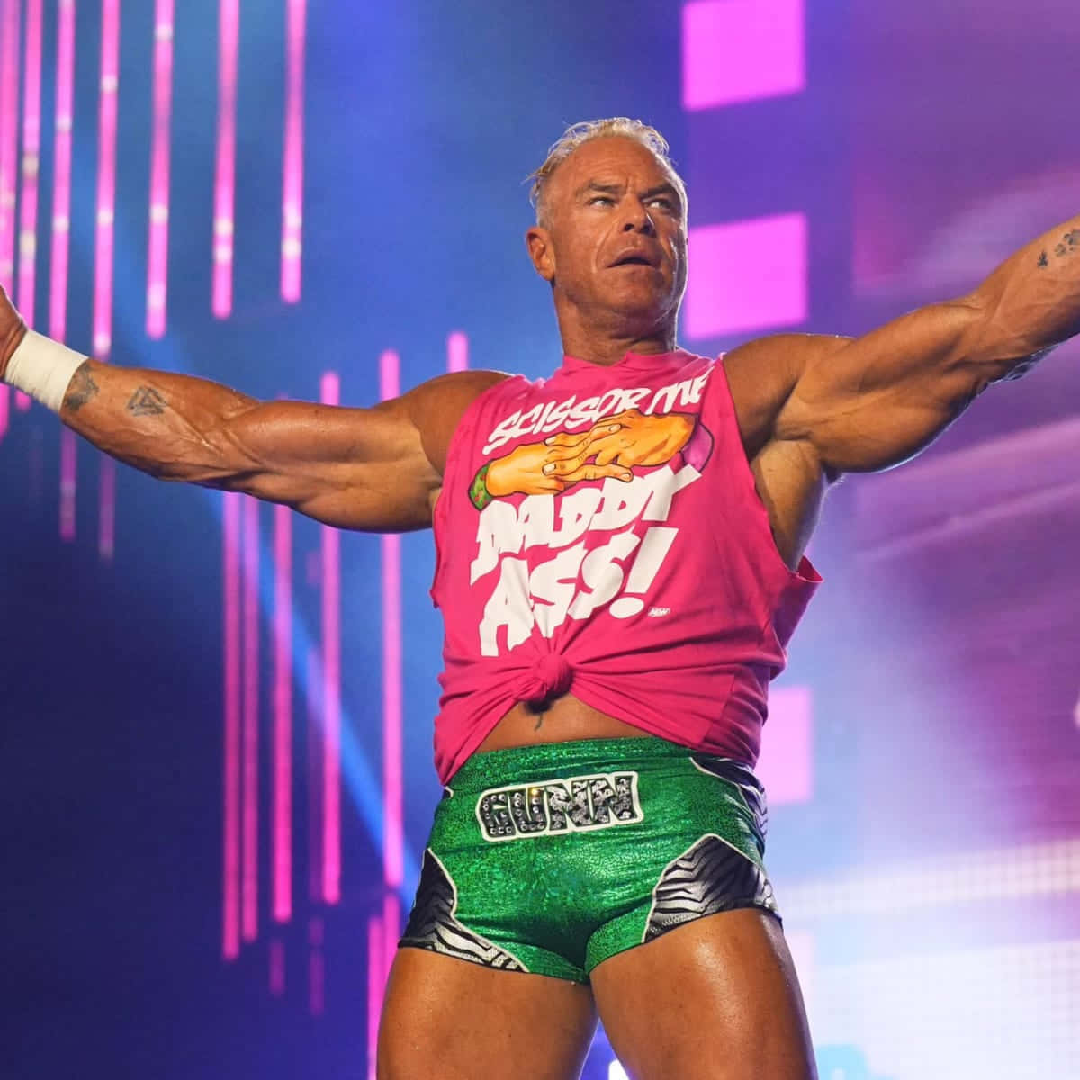 WWE 桌布 壁紙 — 一位肌肉發達的摔角手穿著粉紅色「Scissor Me Daddy Ass!」T恤和綠色短褲，在充滿活力的紫色和藍色舞台燈光下擺出勝利姿勢。