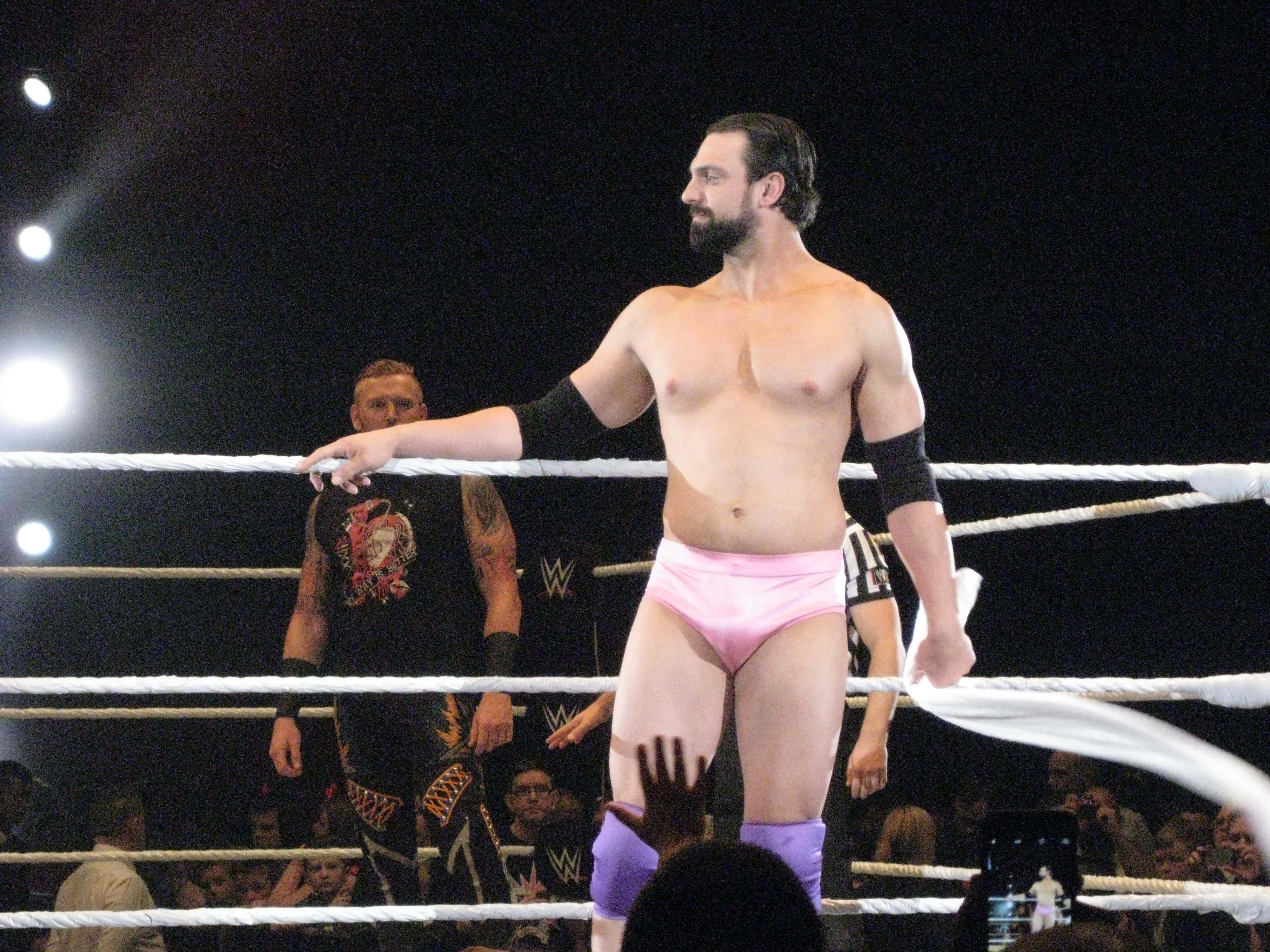 WWE 桌布 壁紙 — 一位肌肉發達的摔角手穿著粉紅色短褲，自信地站在燈光明亮的摔角擂台上，觀眾和裁判清晰可見。