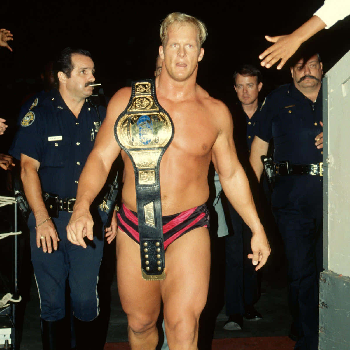 WWE 桌布 壁紙 — 一位肌肉發達的摔角手穿著粉紅色短褲，佩戴金色冠軍腰帶向前邁進，警察在昏暗的環境中護送他。