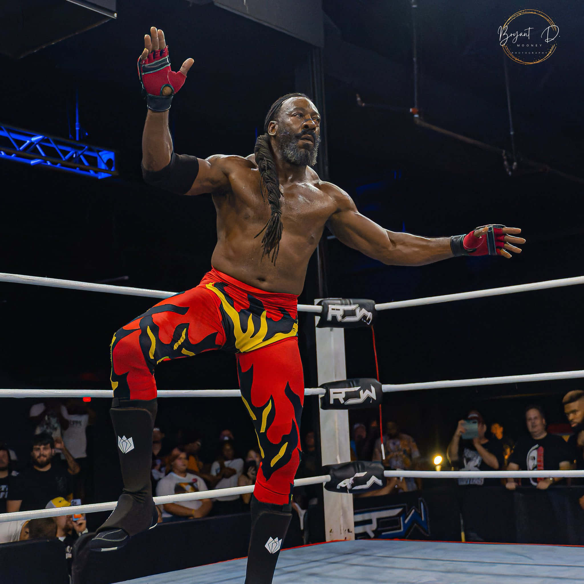 WWE 桌布 壁紙 — 一位肌肉發達的摔角手穿著紅色火焰圖案短褲，在摔角擂台角柱上勝利地站著，在場館燈光下向人群揮手。