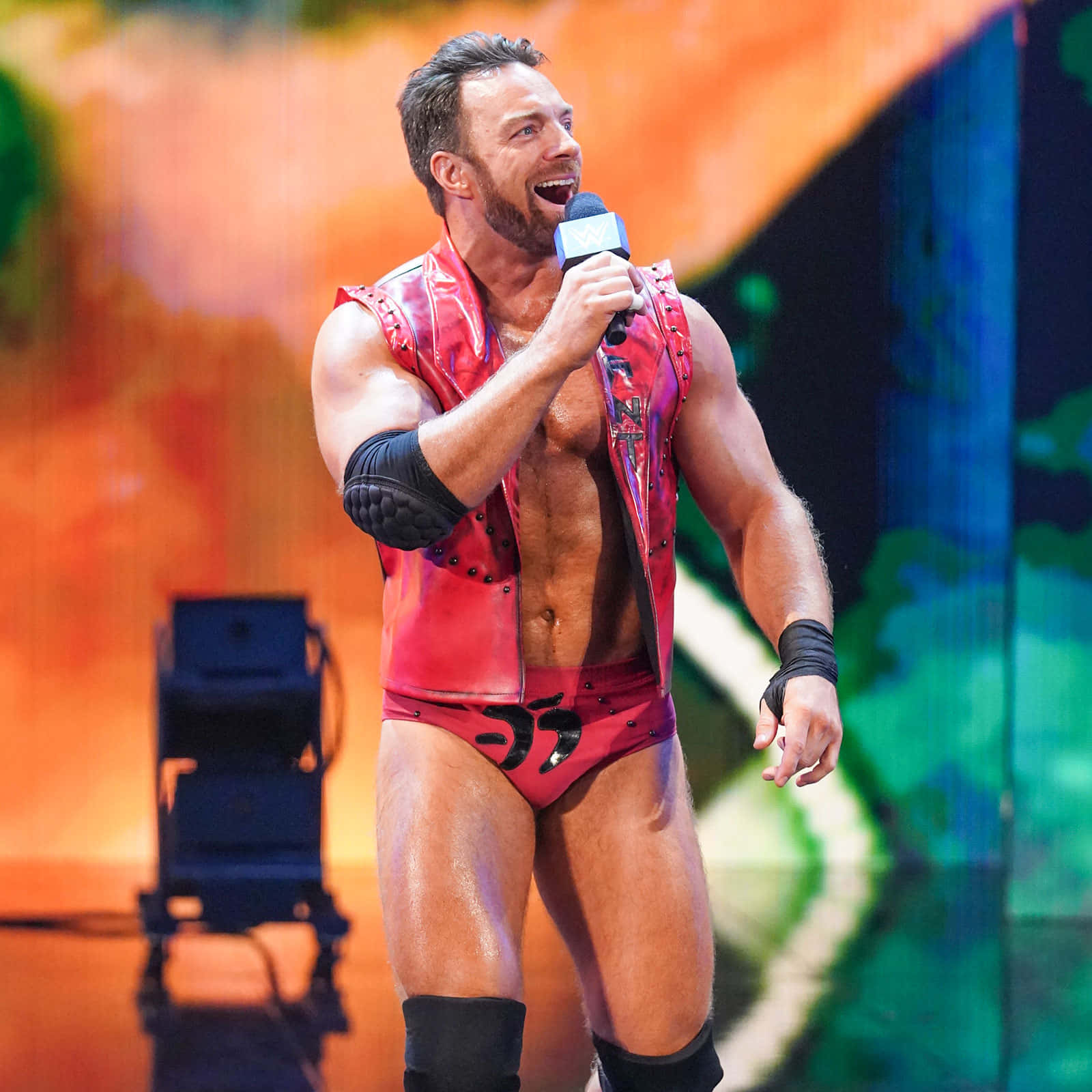 WWE 桌布 壁紙 — 一位肌肉發達的摔角手穿著紅色皮背心和短褲，在明亮的舞台燈光下拿著麥克風激情吶喊。