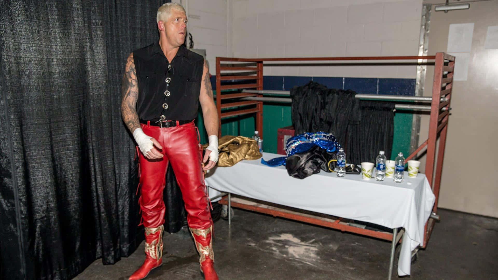 WWE 桌布 壁紙 — 一位肌肉發達的摔角手穿著紅色皮褲和白色手套，站在後台區域，旁邊有一張放有裝備和水瓶的桌子。