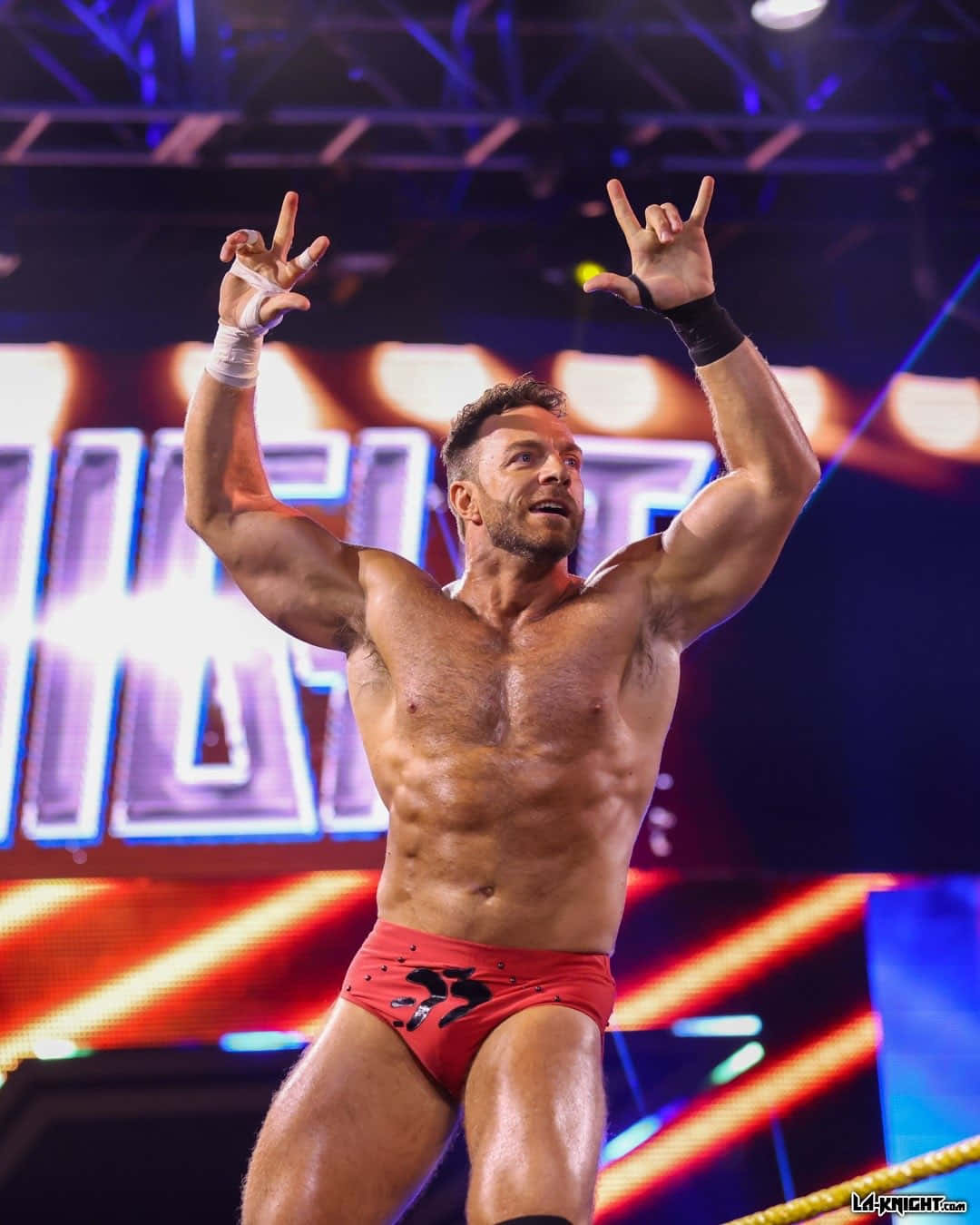 WWE 桌布 壁紙 — 一位肌肉發達的摔角手穿著紅色短褲，在燈光明亮的擂台上，背後有閃耀的舞台燈光，勝利地慶祝。