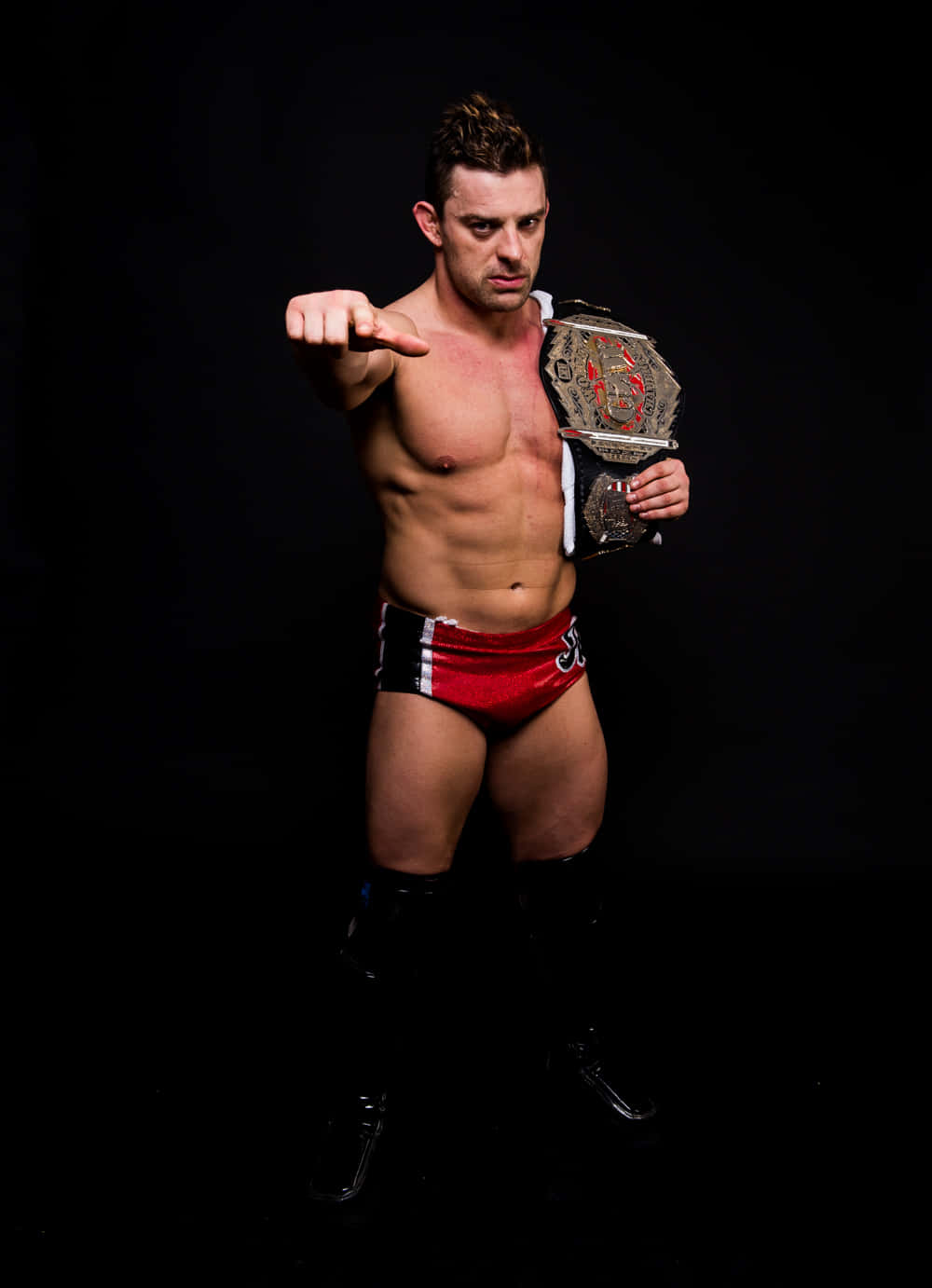 WWE 桌布 壁紙 — 一位肌肉發達的摔角手穿著紅色短褲，手持冠軍腰帶，在黑色背景前直接指向鏡頭。