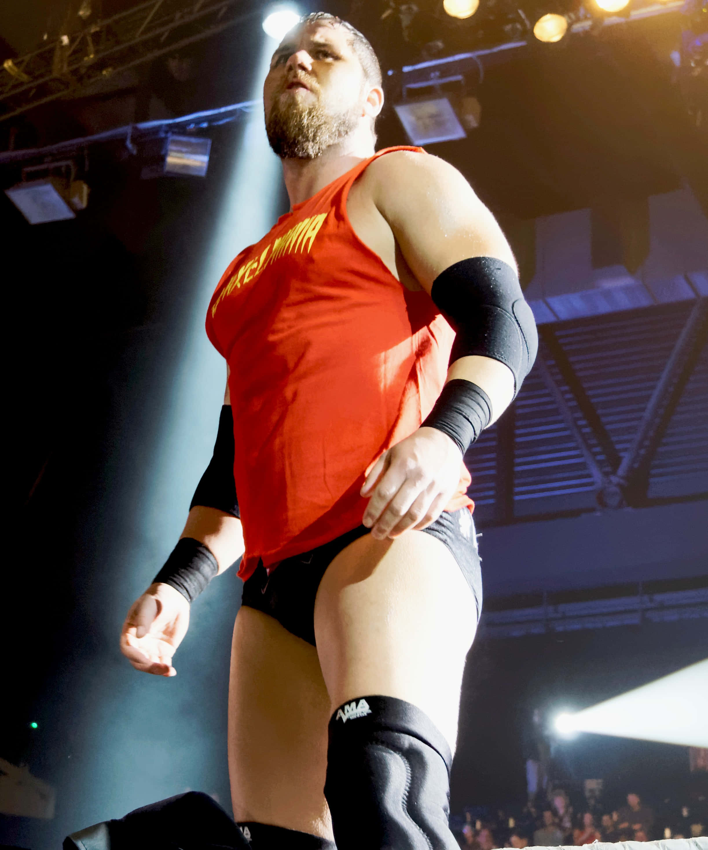 WWE 桌布 壁紙 — 一位肌肉發達的摔角手穿著紅色背心和黑色裝備，在摔角擂台上自信地站在明亮的舞台燈光下。