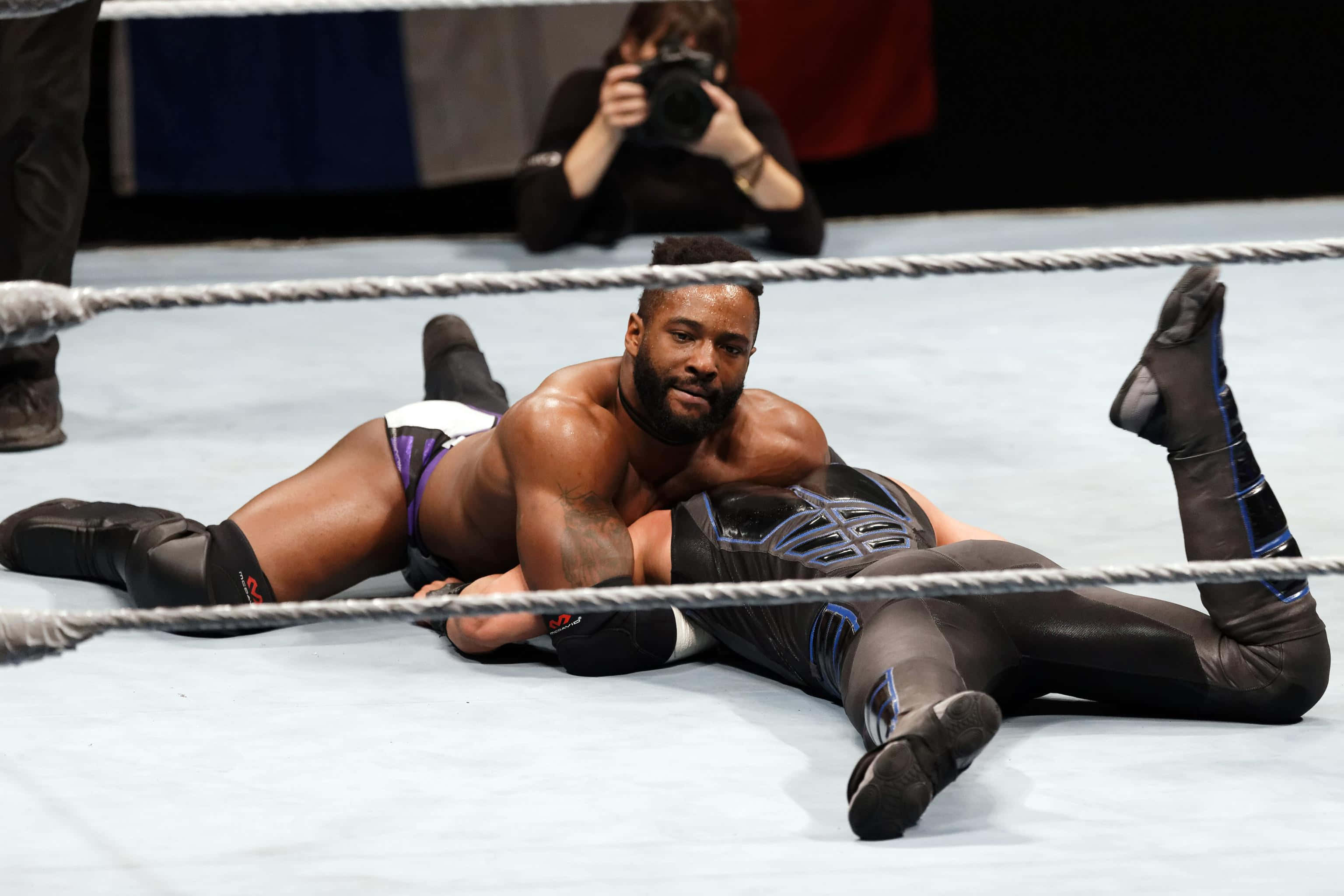 WWE 桌布 壁紙 — 一位肌肉發達的摔角手穿著紫色緊身褲，在明亮的場館燈光下將對手壓制在摔角台上。