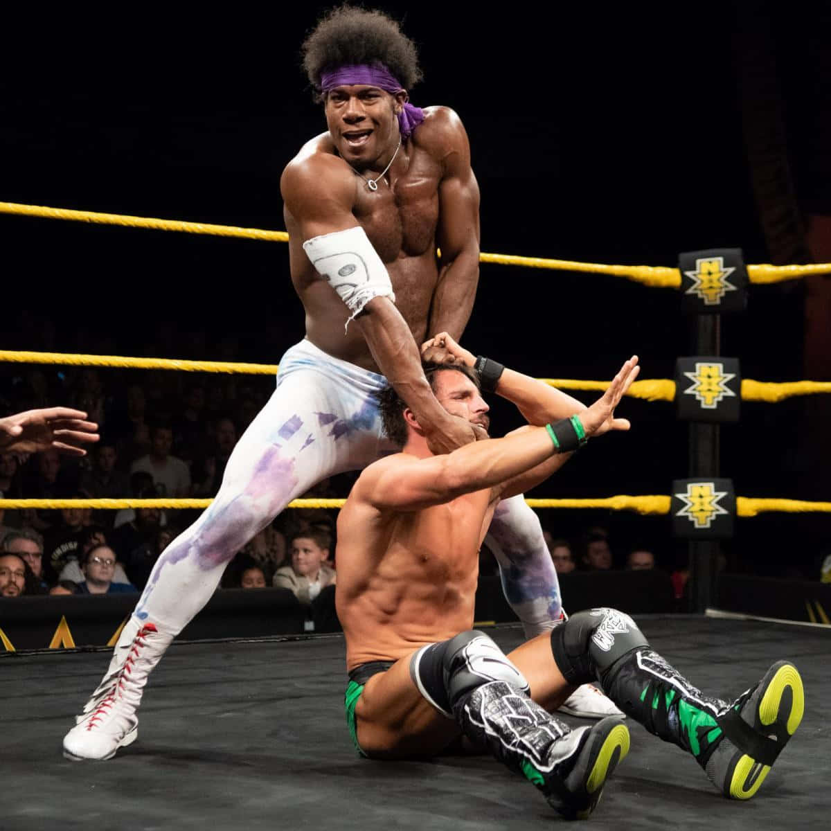 WWE 桌布 壁紙 — 一位肌肉發達的摔角手穿著紮染褲，在燈光明亮的擂台上將對手舉起準備摔擊，周圍有歡呼的粉絲。