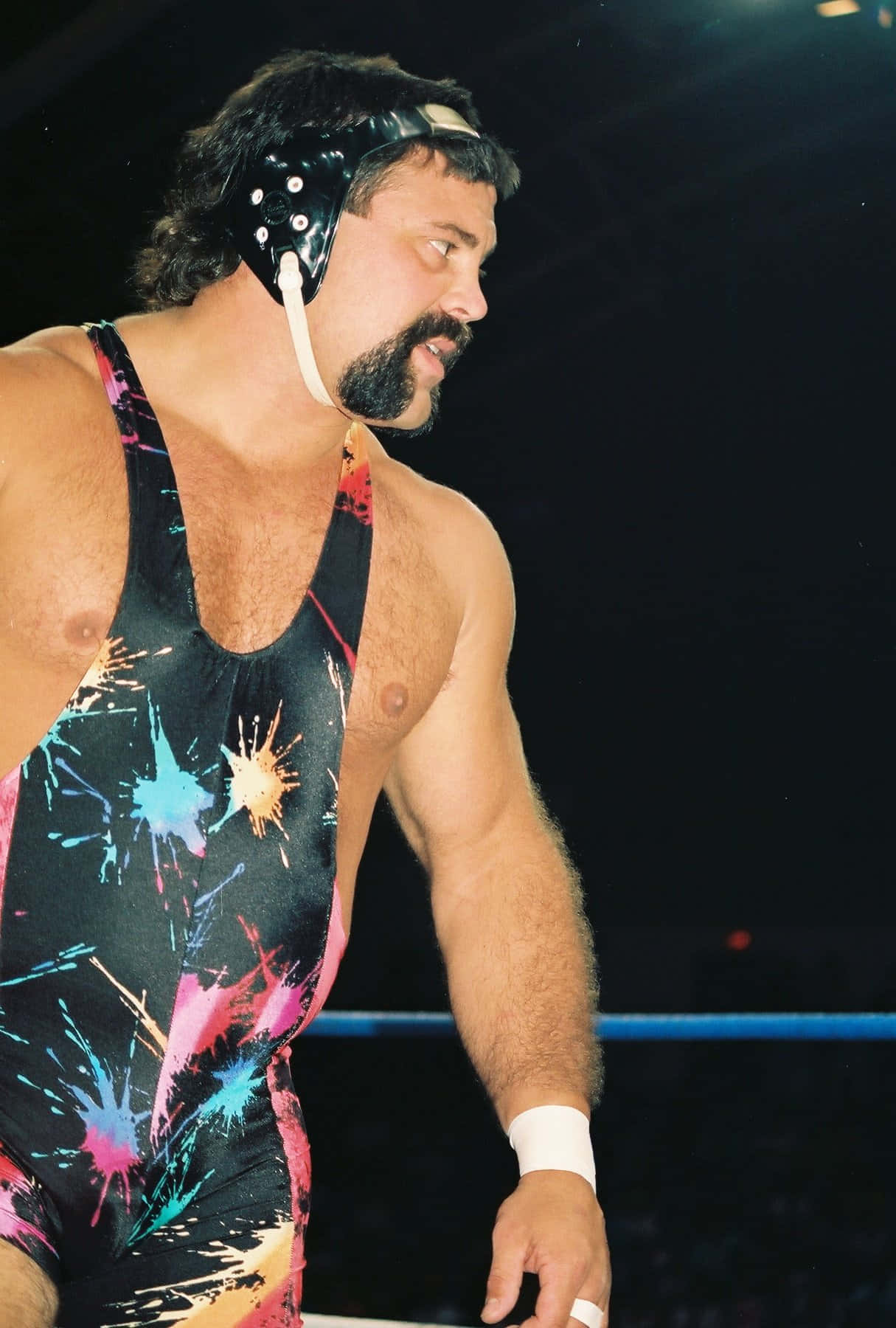 WWE 桌布 壁紙 — 一位肌肉發達的摔角手穿著色彩繽紛的顏料飛濺緊身衣和黑色頭飾，在明亮的競技場燈光下站在擂台上。