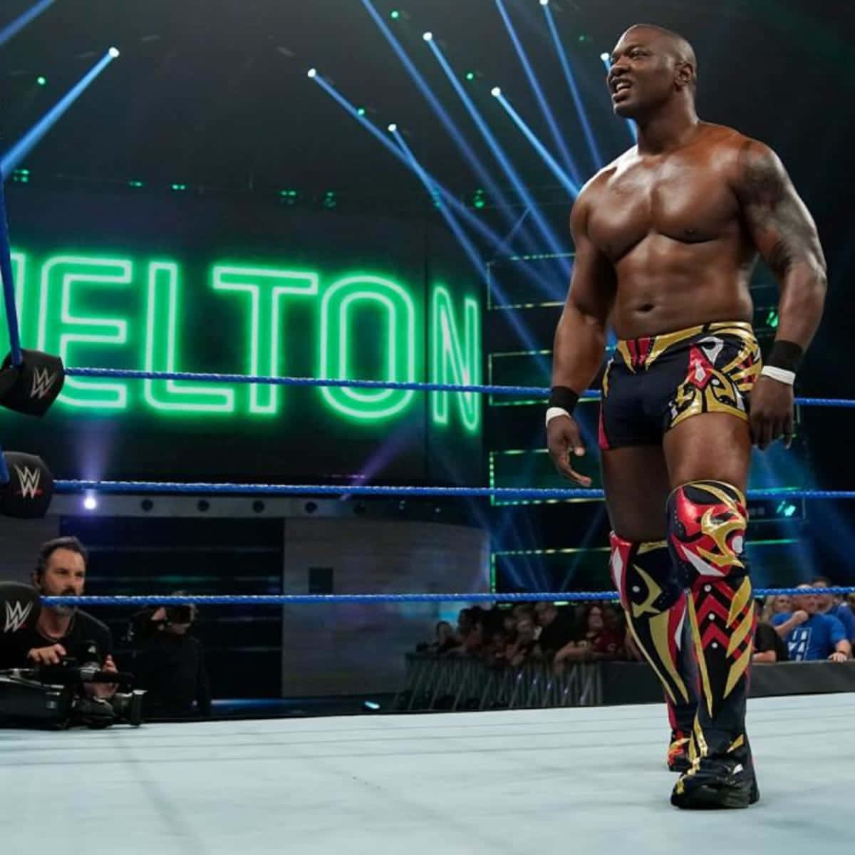 WWE 桌布 壁紙 — 一位肌肉發達的摔角手穿著色彩繽紛的裝備，自信地站在燈光明亮的擂台上，身後『WELTON』發出綠色光芒。