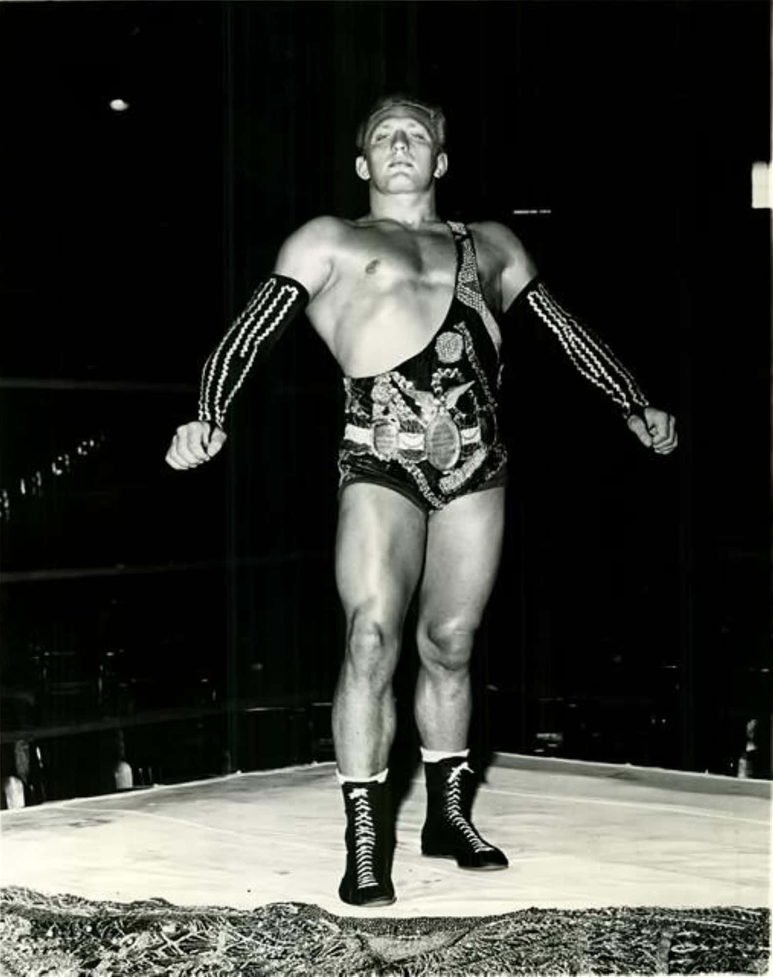 WWE 桌布 壁紙 — 一位肌肉發達的摔角手穿著華麗的短褲和靴子，在戲劇性的黑白燈光下自信地站在擂台上。
