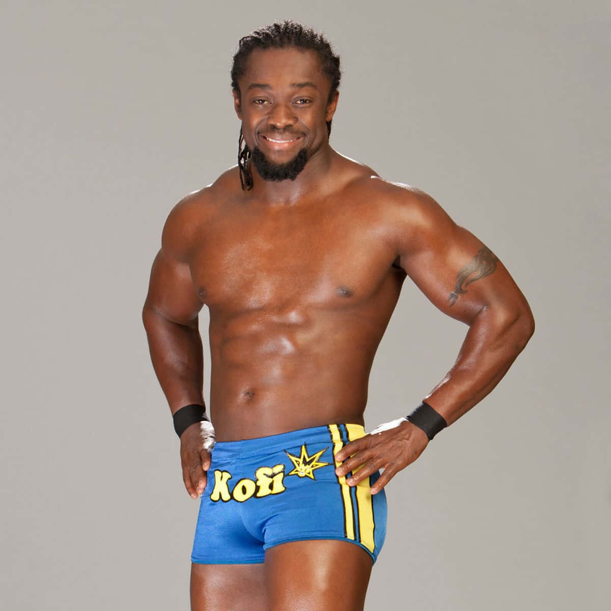 WWE 桌布 壁紙 — 一位肌肉發達的摔角手穿著藍色和黃色短褲，雙手叉腰，在純灰色背景前自信地微笑。