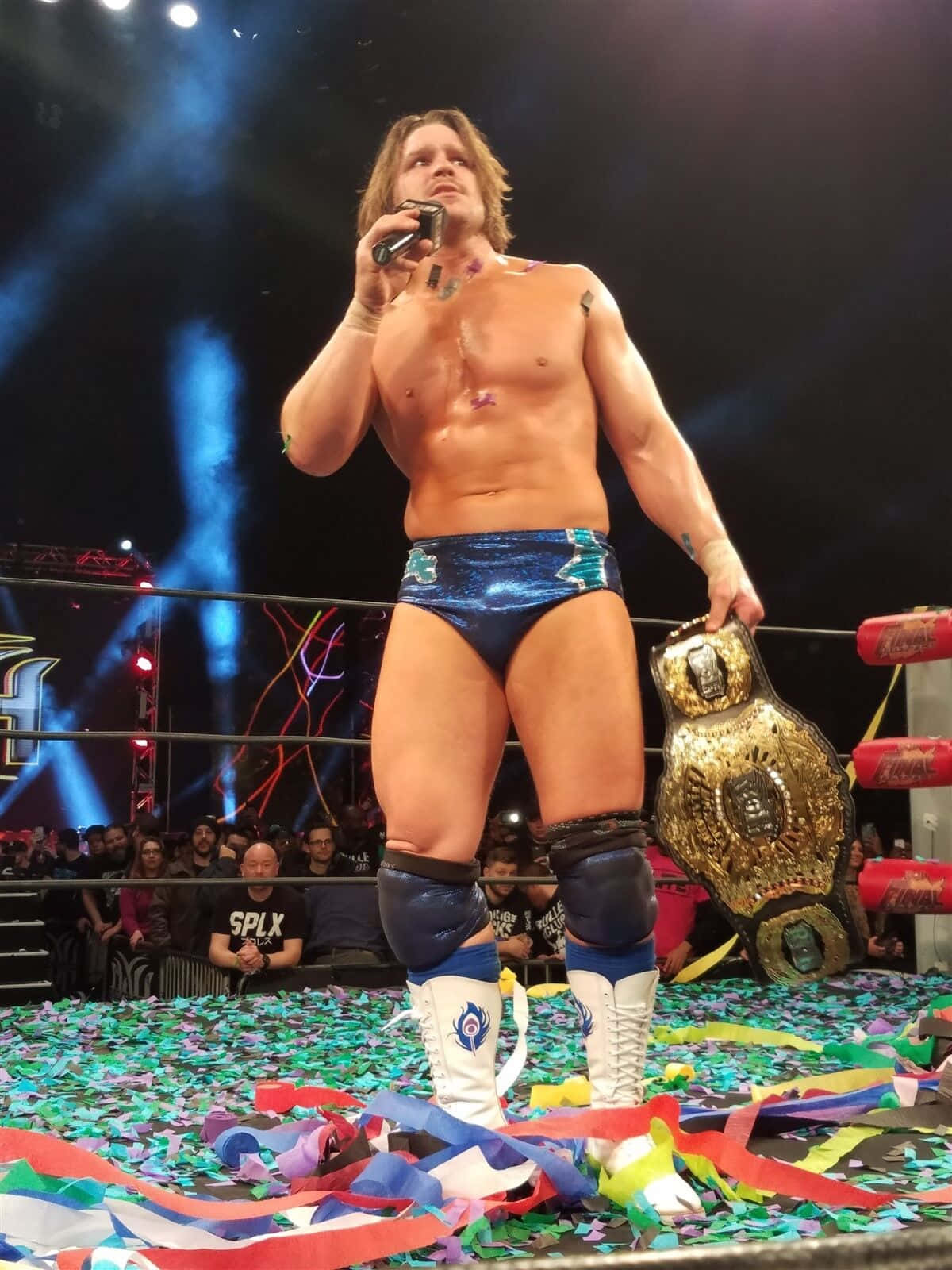 Aew 桌布 壁紙 — 一位肌肉發達的摔角手穿著藍色短褲，在五彩紙屑中手持冠軍腰帶和麥克風，在擂台上勝利地站立。