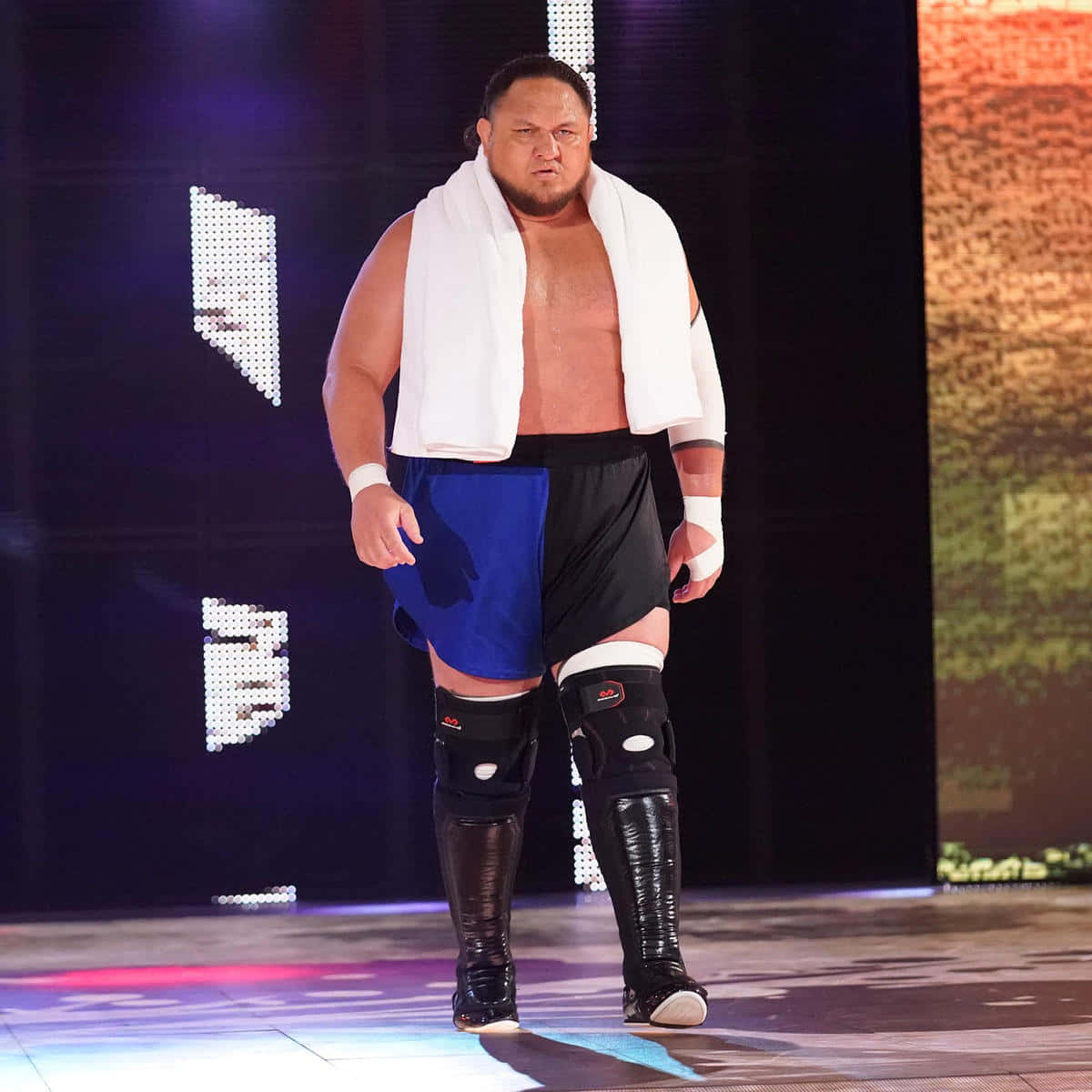 WWE 桌布 壁紙 — 一位肌肉發達的摔角手穿著藍黑短褲，肩上披著白色毛巾，自信地走過燈光明亮的擂台入口。