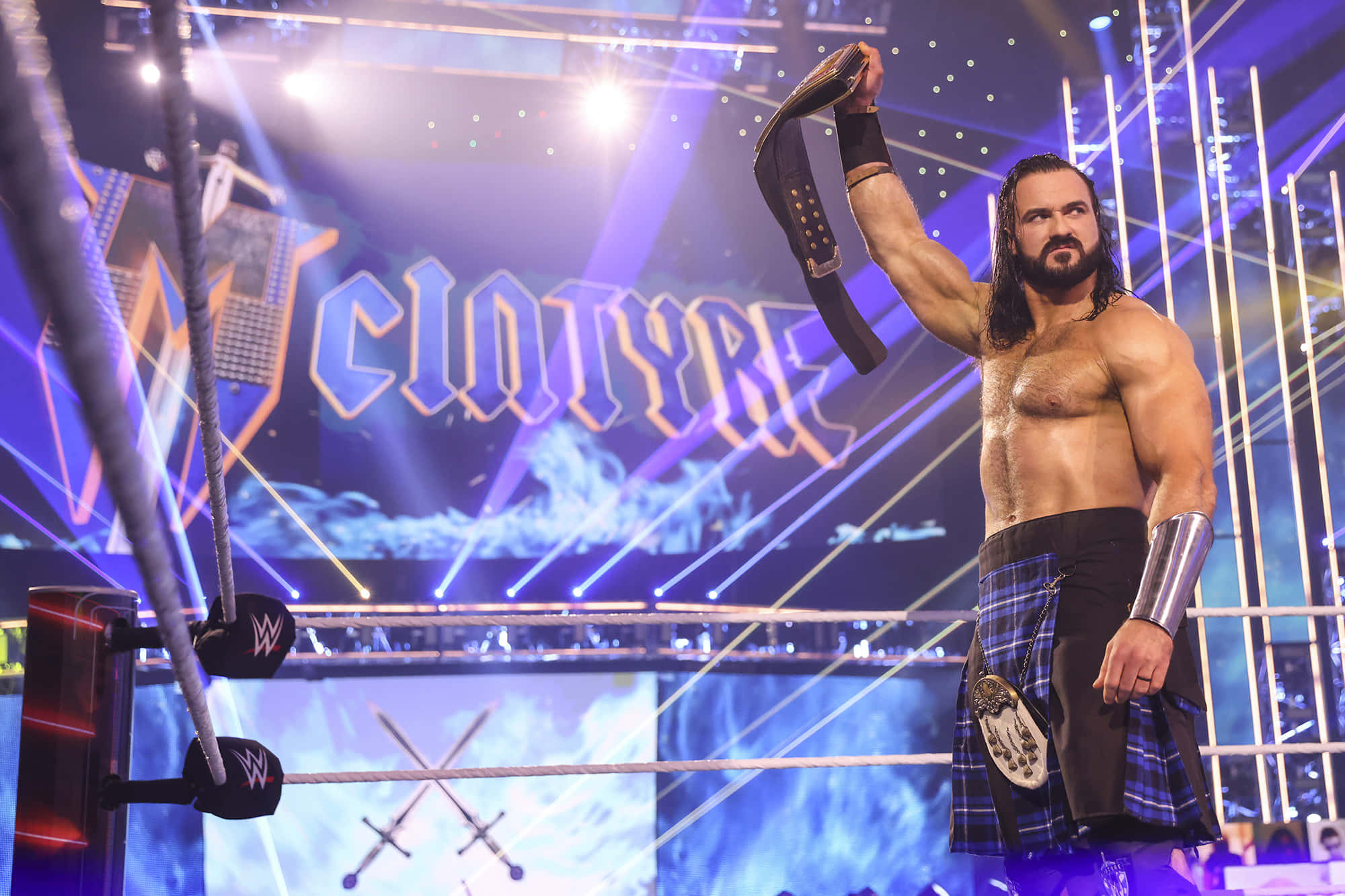 WWE 桌布 壁紙 — 一位肌肉發達的摔角手穿著蘇格蘭裙，在燈光明亮的摔角場中勝利地高舉冠軍腰帶。