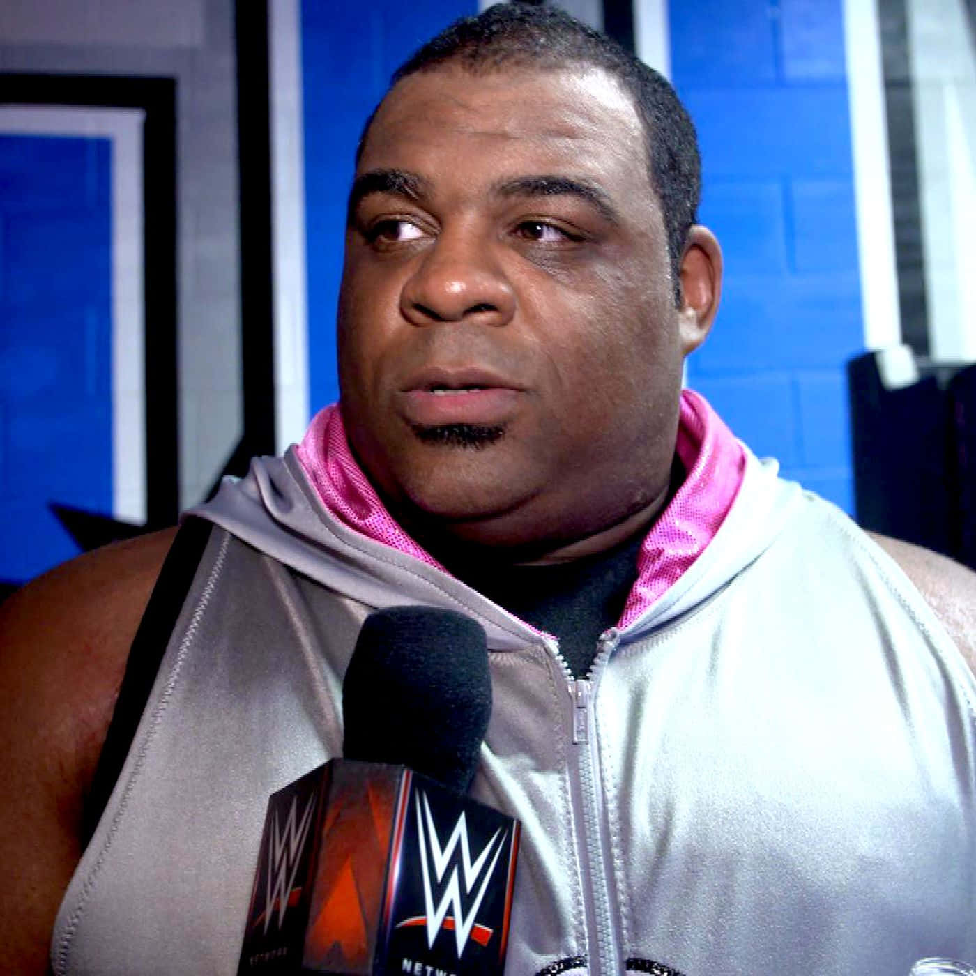 WWE 桌布 壁紙 — 一位肌肉發達的摔角手穿著銀色連帽衫，內襯粉紅色，對著WWE Network麥克風說話，背景是藍色磚牆。