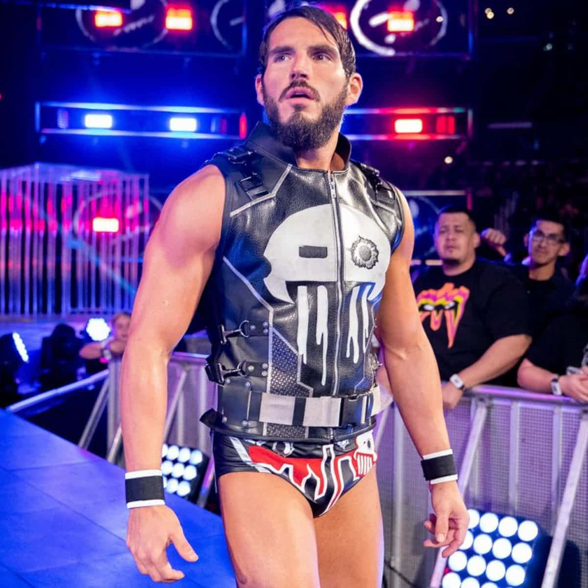 WWE 桌布 壁紙 — 一位肌肉發達的摔角手穿著骷髏主題皮背心，自信地走過燈光明亮的競技場，背景有發光的舞台燈光和歡呼的粉絲。