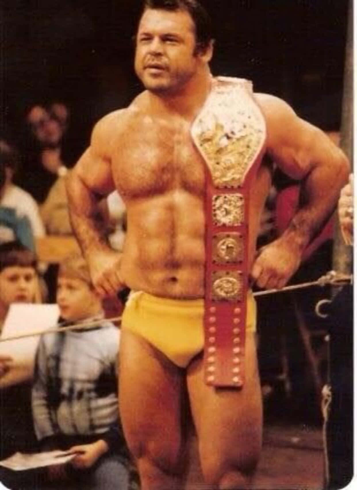 Ivan Putski 桌布 壁紙 — 一位肌肉發達的摔角手穿著黃色短褲，自信地站在擂台上，佩戴著冠軍腰帶。
