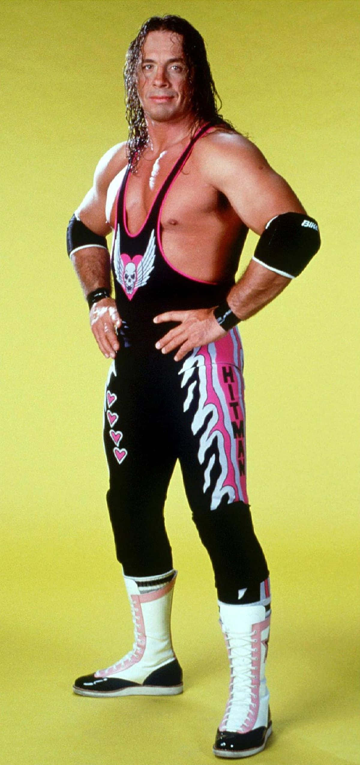 WWE 桌布 壁紙 — 一位肌肉發達的摔角手穿著黑色、粉紅色和白色的服裝，在純黃色背景前自信地擺姿勢。