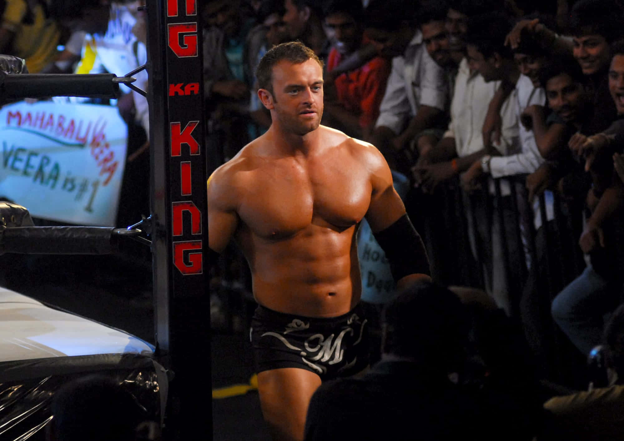 Nick Aldis 桌布 壁紙 — 一位肌肉發達的摔角手穿著黑色三角褲，自信地走過擂台入口，背景是歡呼的粉絲。