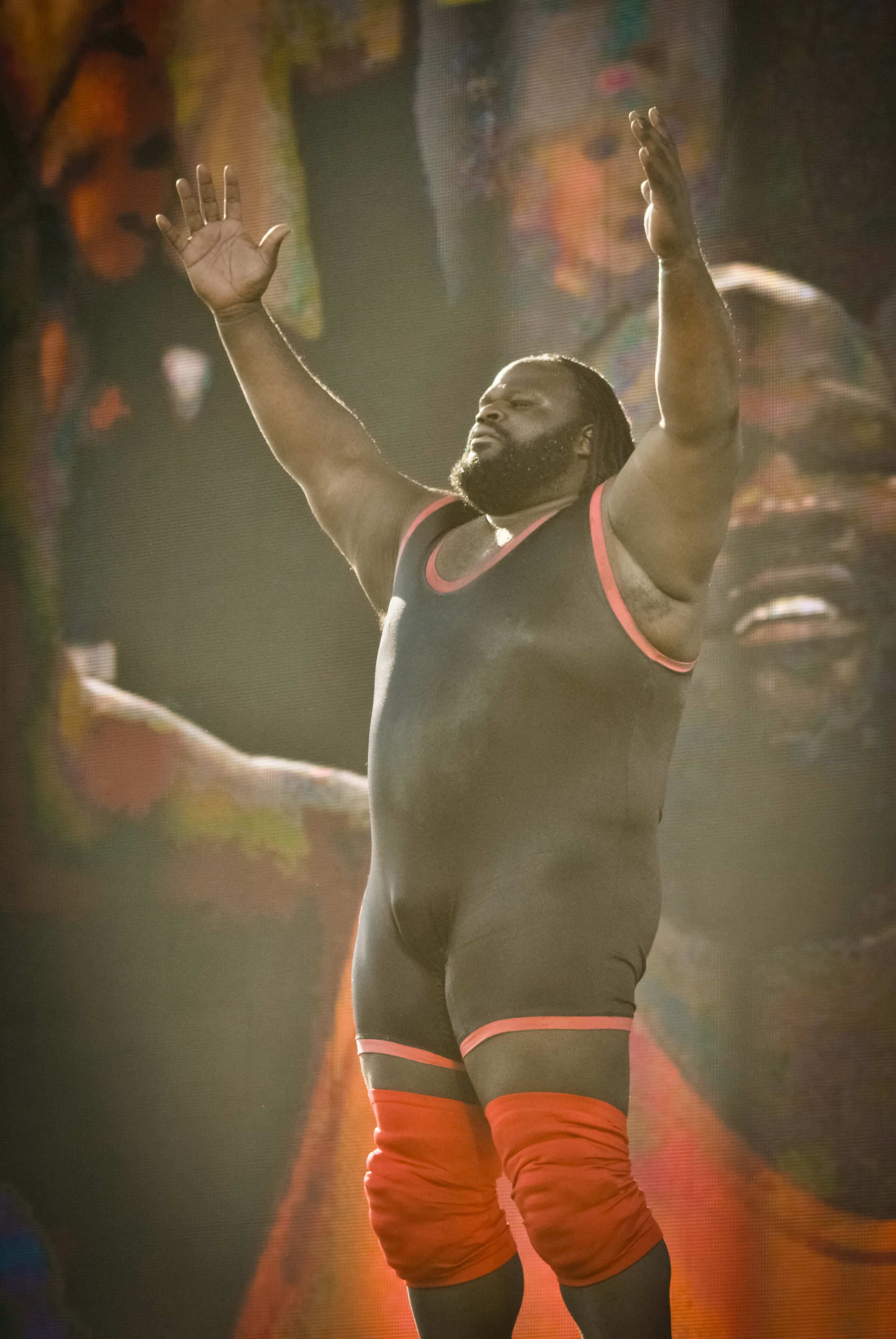 WWE 桌布 壁紙 — 一位肌肉發達的摔角手穿著黑色和橙色裝備，在明亮的舞台燈光下勝利地高舉雙臂。