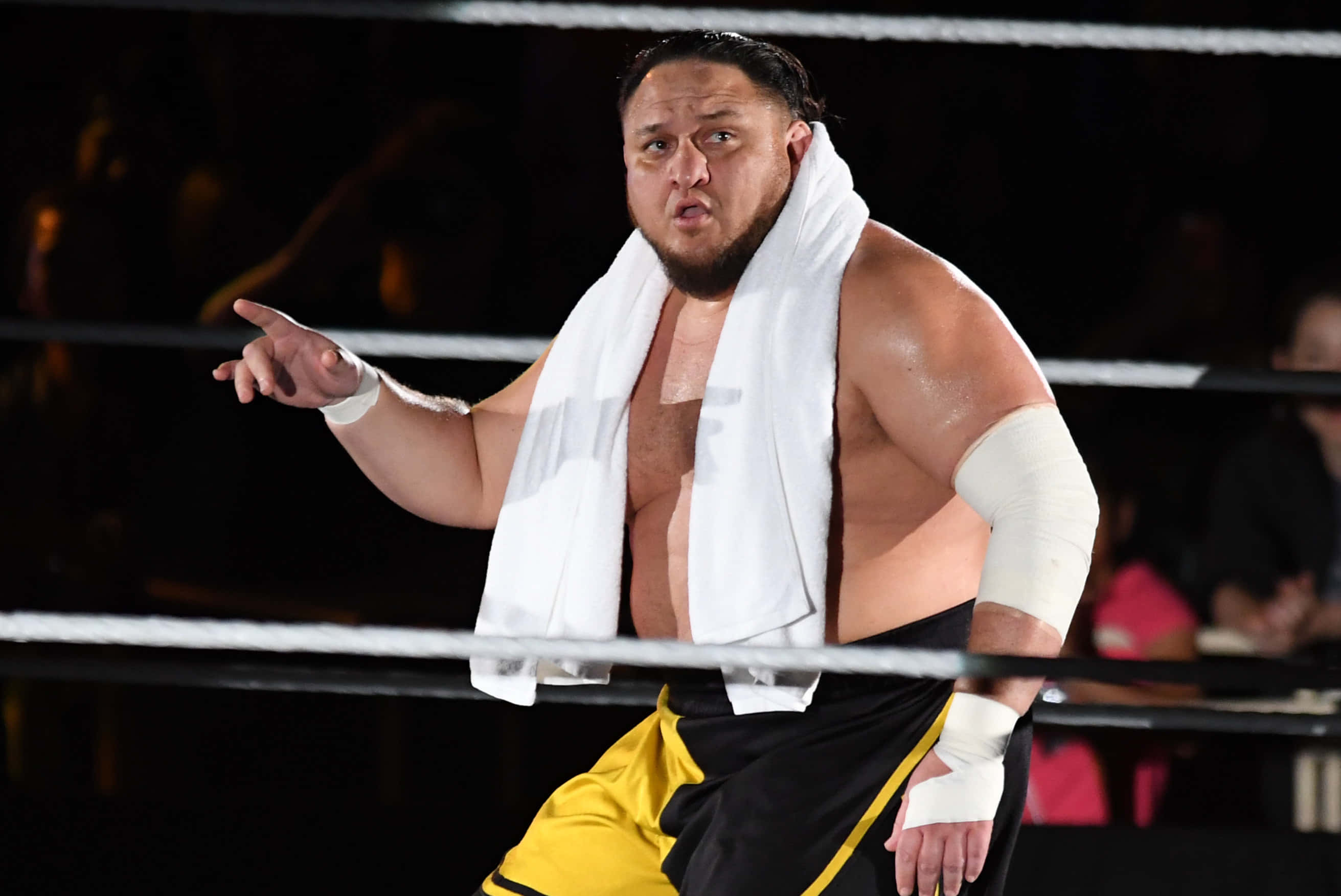 WWE 桌布 壁紙 — 一位肌肉發達的摔角手穿著黑色和黃色短褲，肩上披著白色毛巾，在明亮的擂台上激烈地指著。