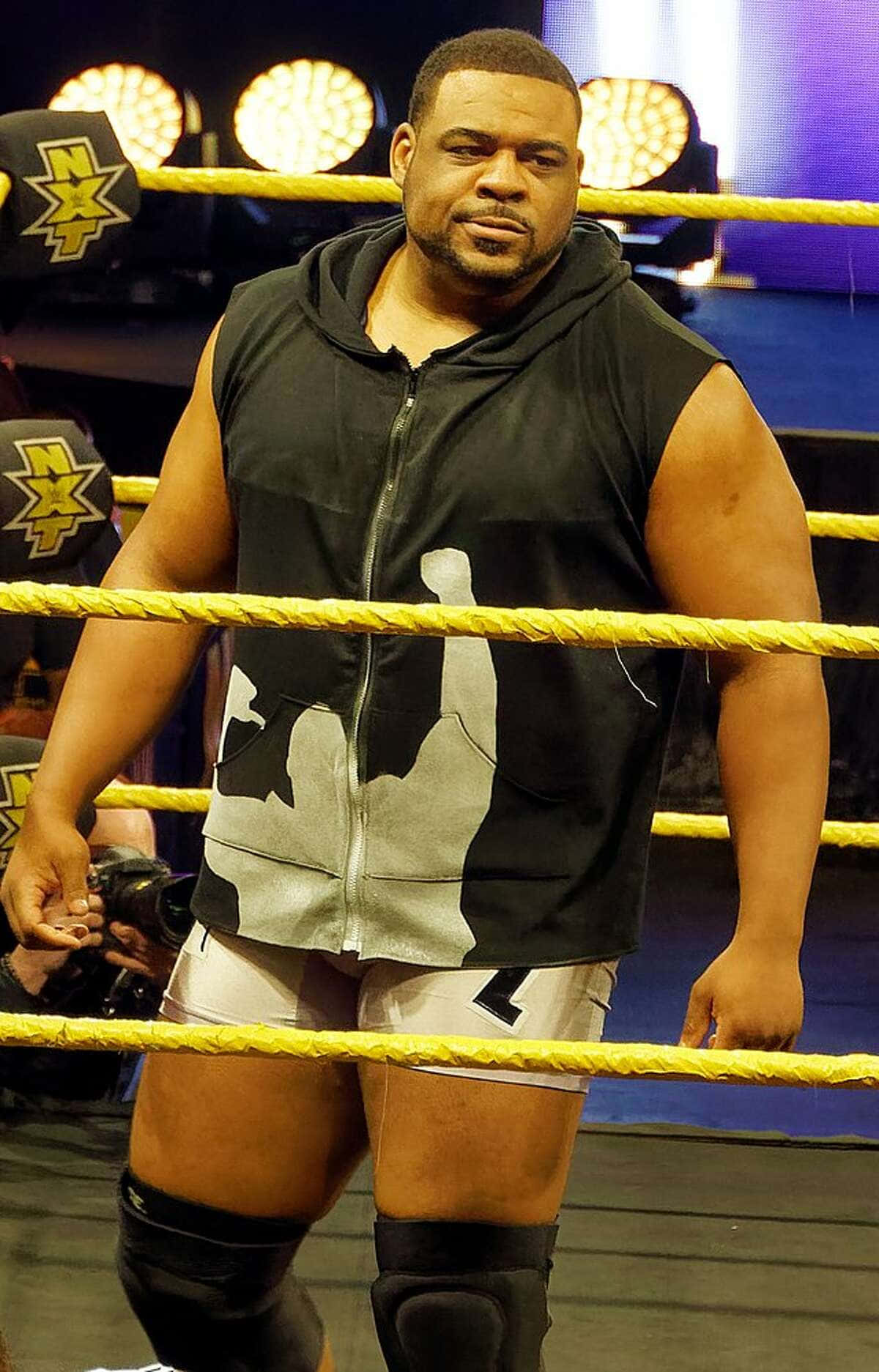 WWE 桌布 壁紙 — 一位肌肉發達的摔角手穿著黑色無袖連帽衫和白色短褲，自信地站在燈光明亮的摔角擂台內，擂台有黃色繩索。