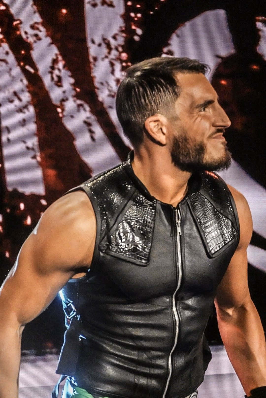 WWE 桌布 壁紙 — 一位肌肉發達的摔角手穿著黑色皮背心，在明亮的舞台燈光下自信地微笑。