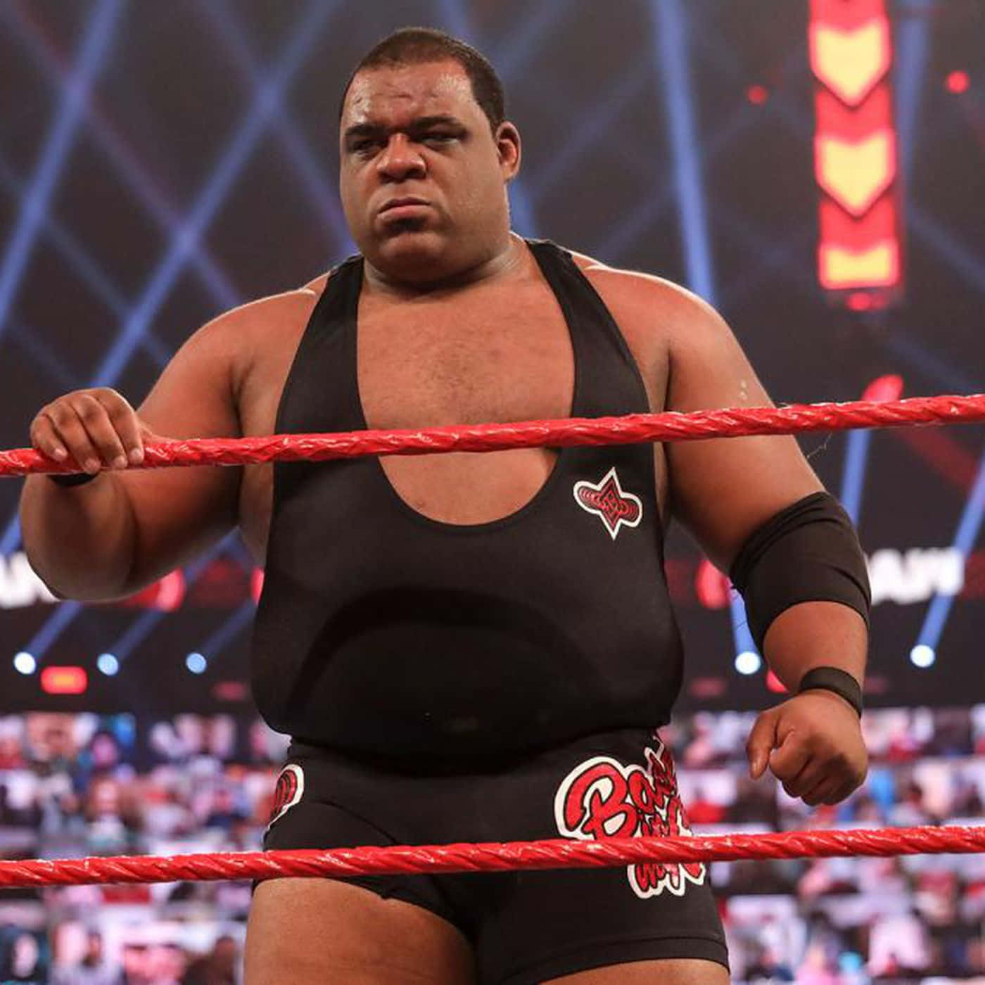 WWE 桌布 壁紙 — 一位肌肉發達的摔角手穿著黑色緊身衣，在明亮的場館燈光下抓住摔角場的紅色繩索。