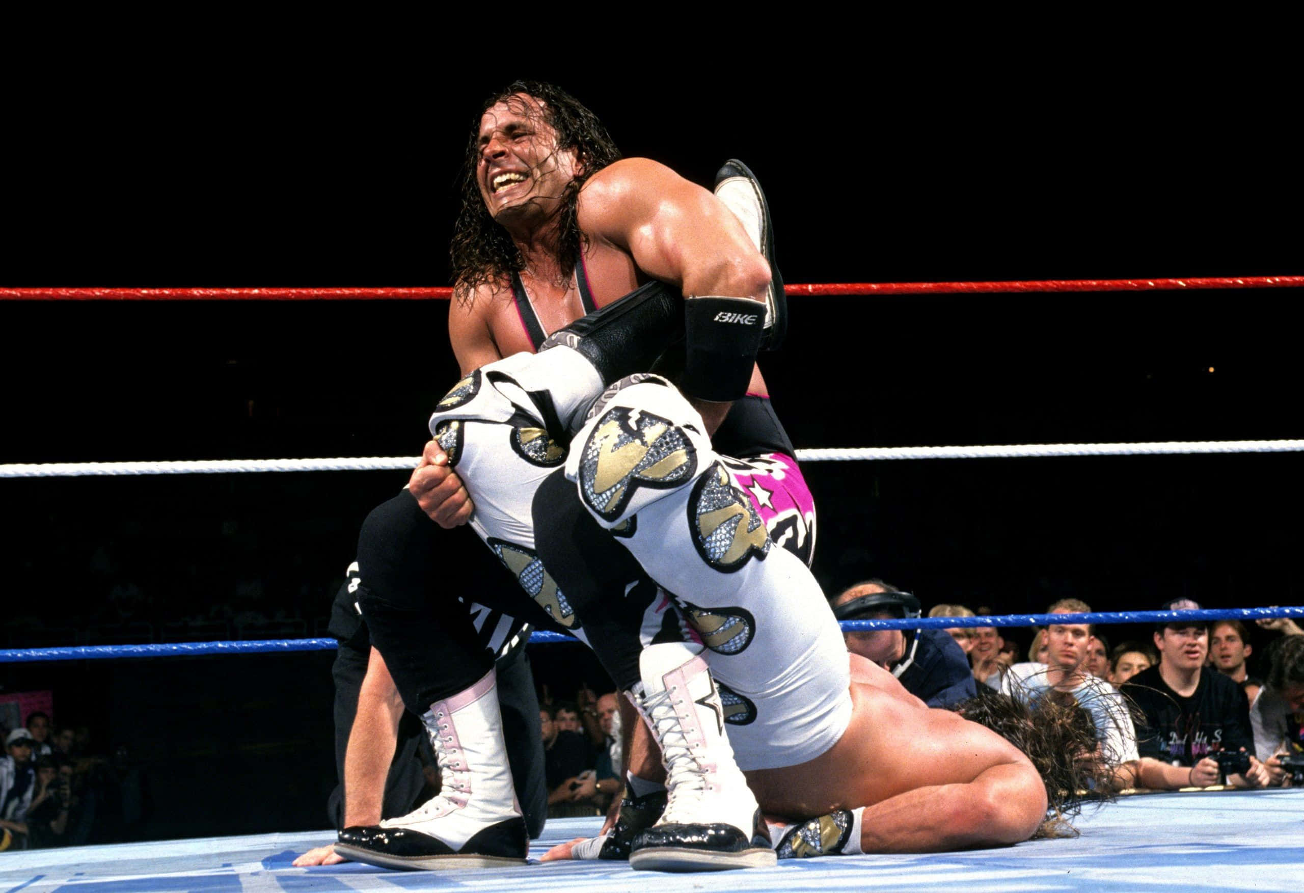 shawn michaels 桌布 壁紙 — 一位肌肉發達的摔角手穿著黑色緊身褲，在明亮的場館燈光下咧嘴笑著，將對手舉起在摔角台上。