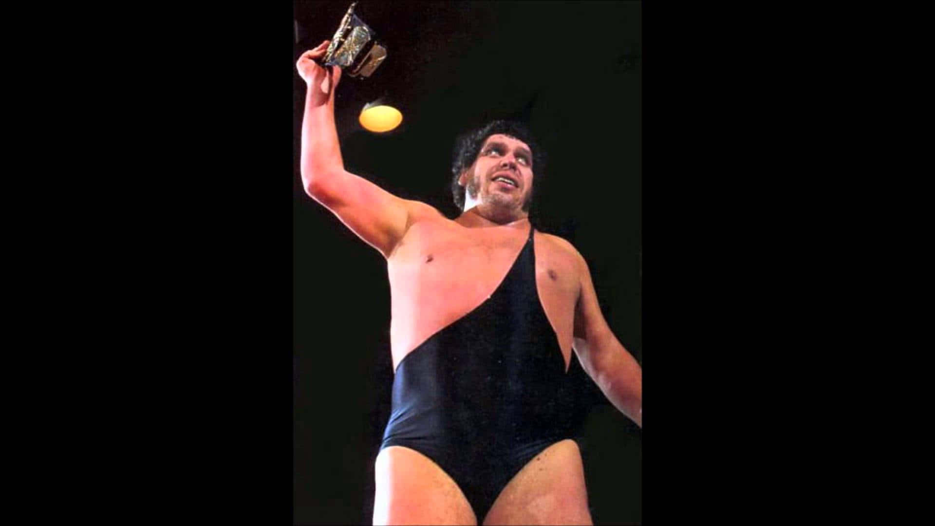 Andre The Giant 桌布 壁紙 — 一位肌肉發達的摔角手穿著黑色緊身衣，在明亮的舞台燈光下勝利地高舉冠軍腰帶。