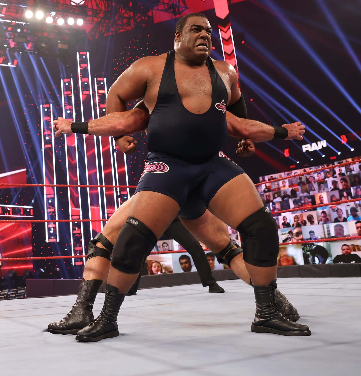 WWE 桌布 壁紙 — 一位肌肉發達的摔角手穿著黑色緊身衣，在燈光明亮的擂台上咆哮，背後螢幕上有紅藍燈光和粉絲人群。