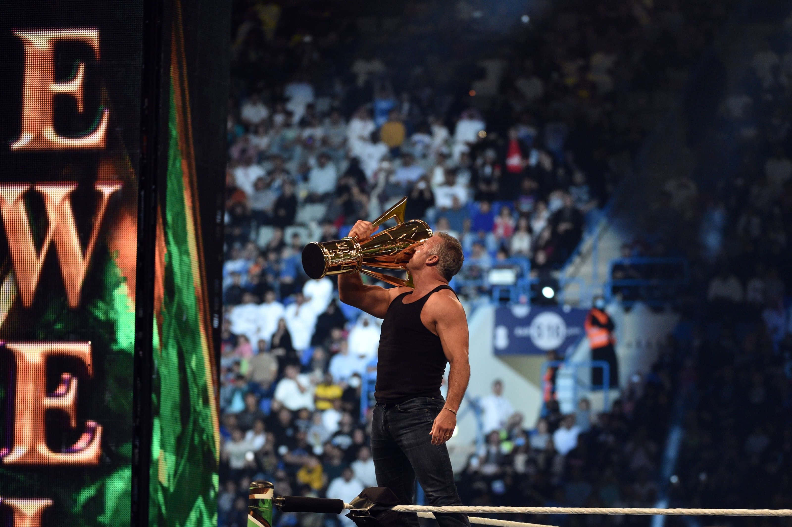 Wrestling 桌布 壁紙 — 一位肌肉發達的摔角手穿著黑色背心，在摔角場內親吻一個大型金色獎盃，身後有歡呼的人群。