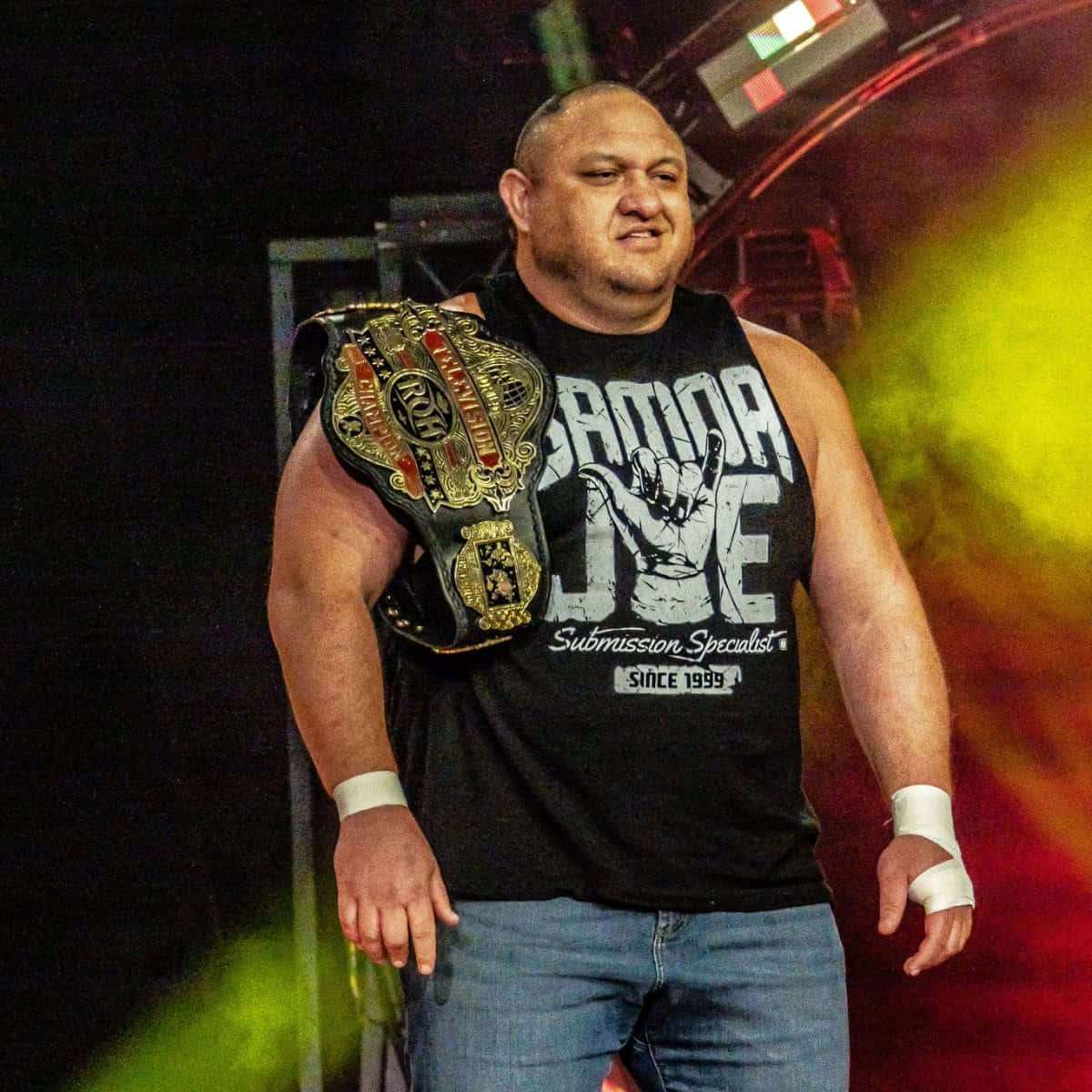 WWE 桌布 壁紙 — 一位肌肉發達的摔角手穿著黑色背心和牛仔褲，在舞台燈光下自豪地展示冠軍腰帶。