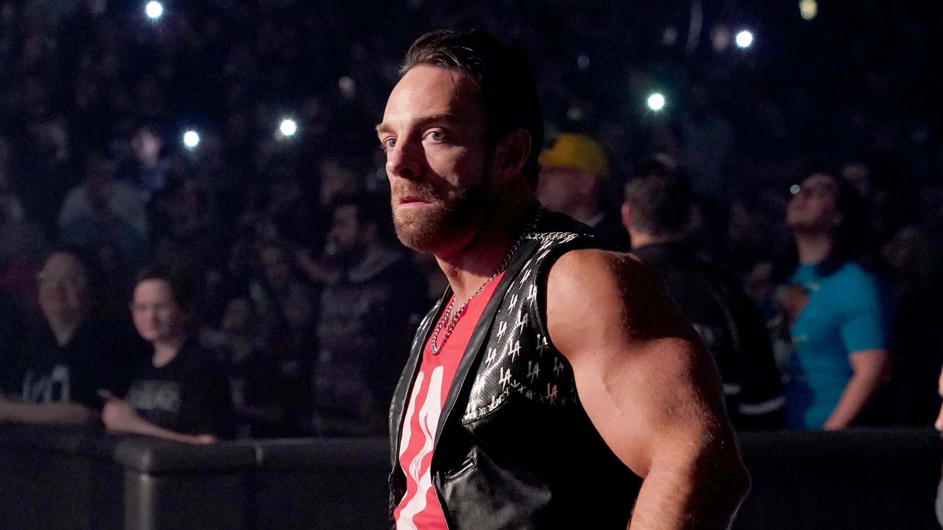 WWE 桌布 壁紙 — 一位肌肉發達的摔角手穿著黑色背心和紅色襯衫，在明亮的聚光燈下自信地站在黑暗的競技場人群前。