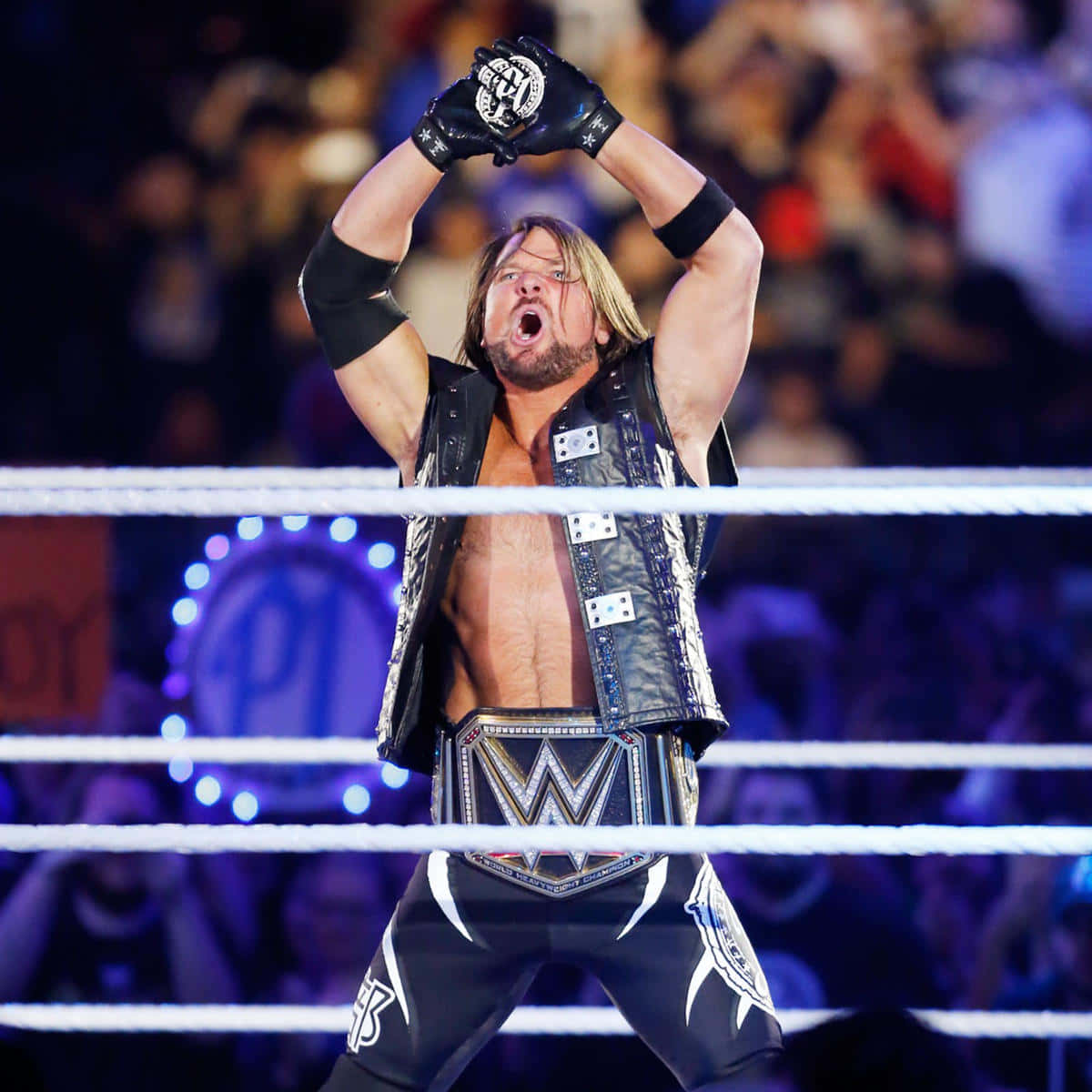 WWE 桌布 壁紙 — 一位肌肉發達的摔角手穿著黑色背心和冠軍腰帶，在燈光明亮的摔角場內舉起戴手套的拳頭慶祝勝利。